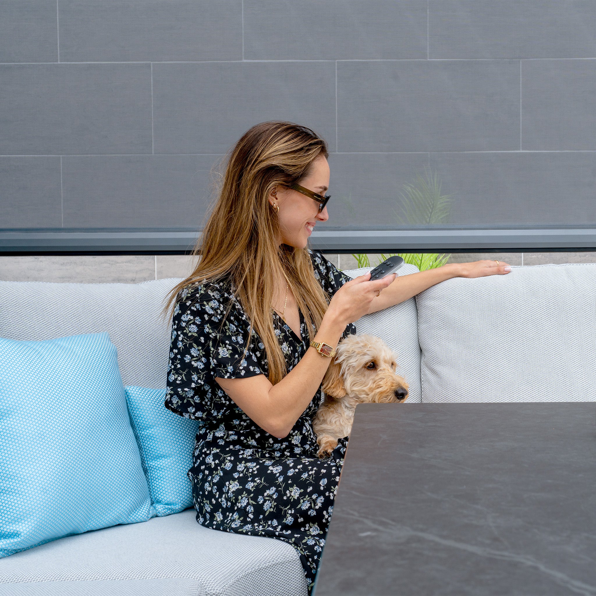A woman in a floral dress sits on a couch with her small dog, holding a phone and smiling under the sleek PergoSTET Deluxe Pergola. Its motorised LED louvres and four manual side blinds create an inviting outdoor setting.