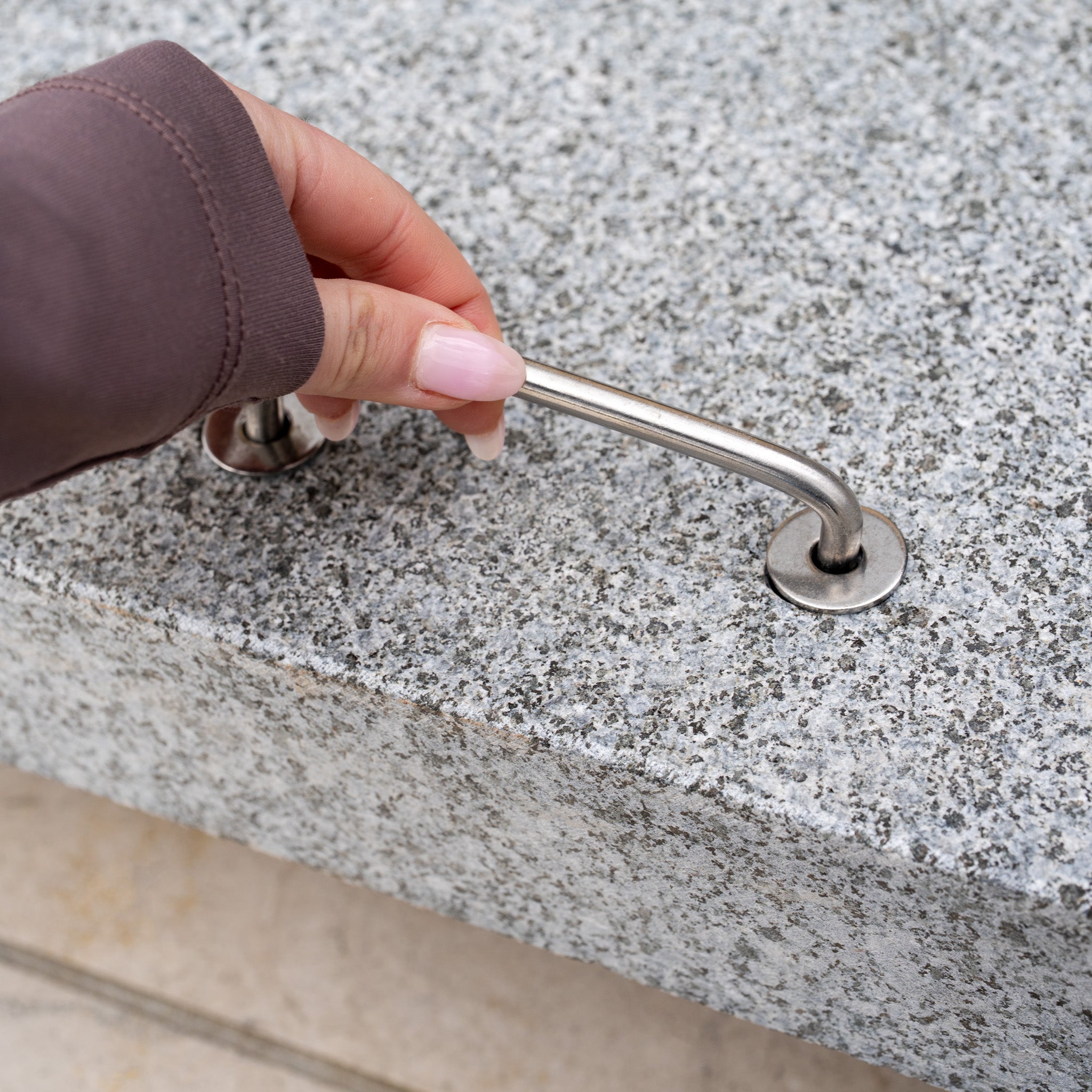 A hand grips the metal handle of the Siesta 125kg Granite Base, a gray granite parasol base ideal for supporting cantilever designs.