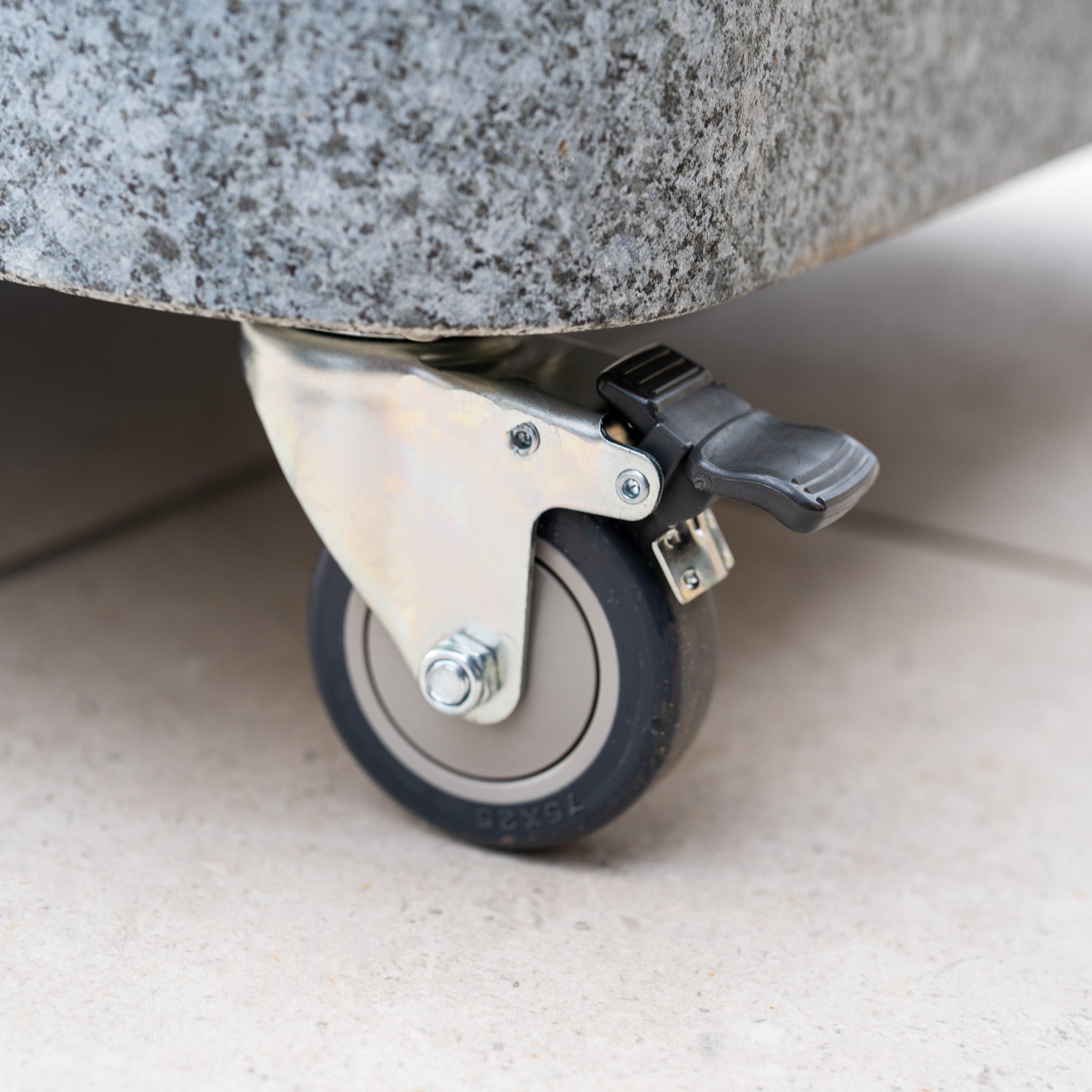 A close-up of a caster wheel with brake attached to the Siesta 125kg Granite Base on a tiled floor—perfect for supporting sturdy outdoor parasol or cantilever designs.