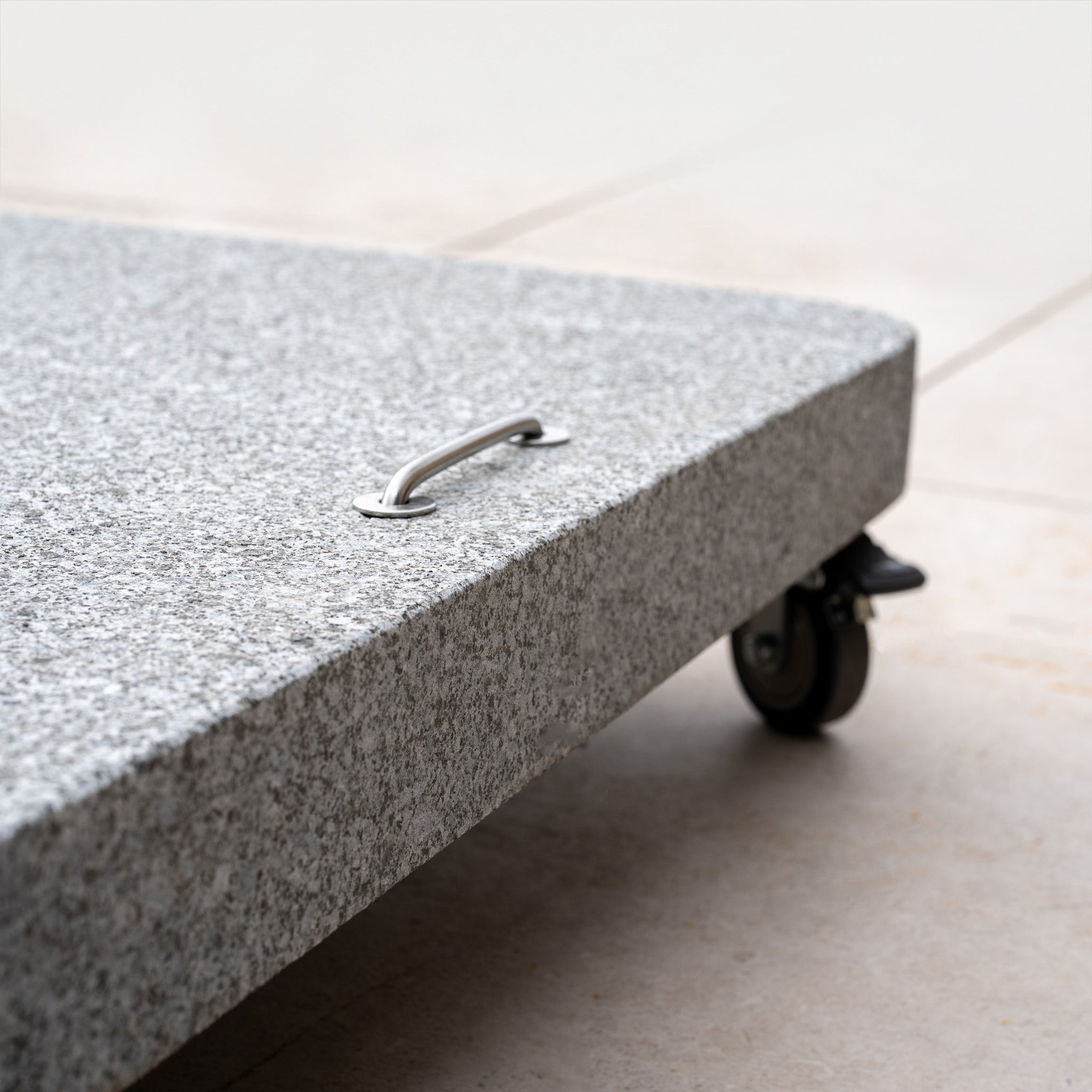 A close-up of the Siesta 125kg Granite Base in gray, featuring a metal handle and caster wheels on a tiled floor—ideal support for outdoor parasols or cantilever designs.
