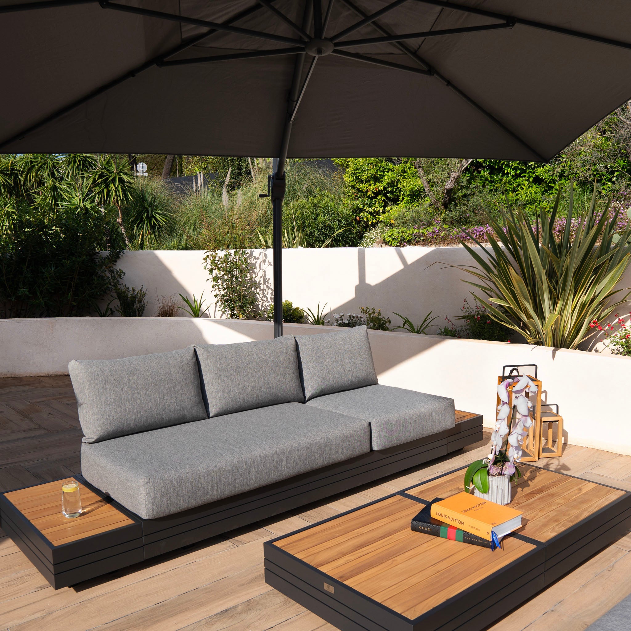 Outdoor patio with a gray sectional sofa under a large Hacienda 3m x 4m Cantilever Parasol with Granite Base & Cover in Charcoal, along with books, lanterns, and a drink on wooden tables.
