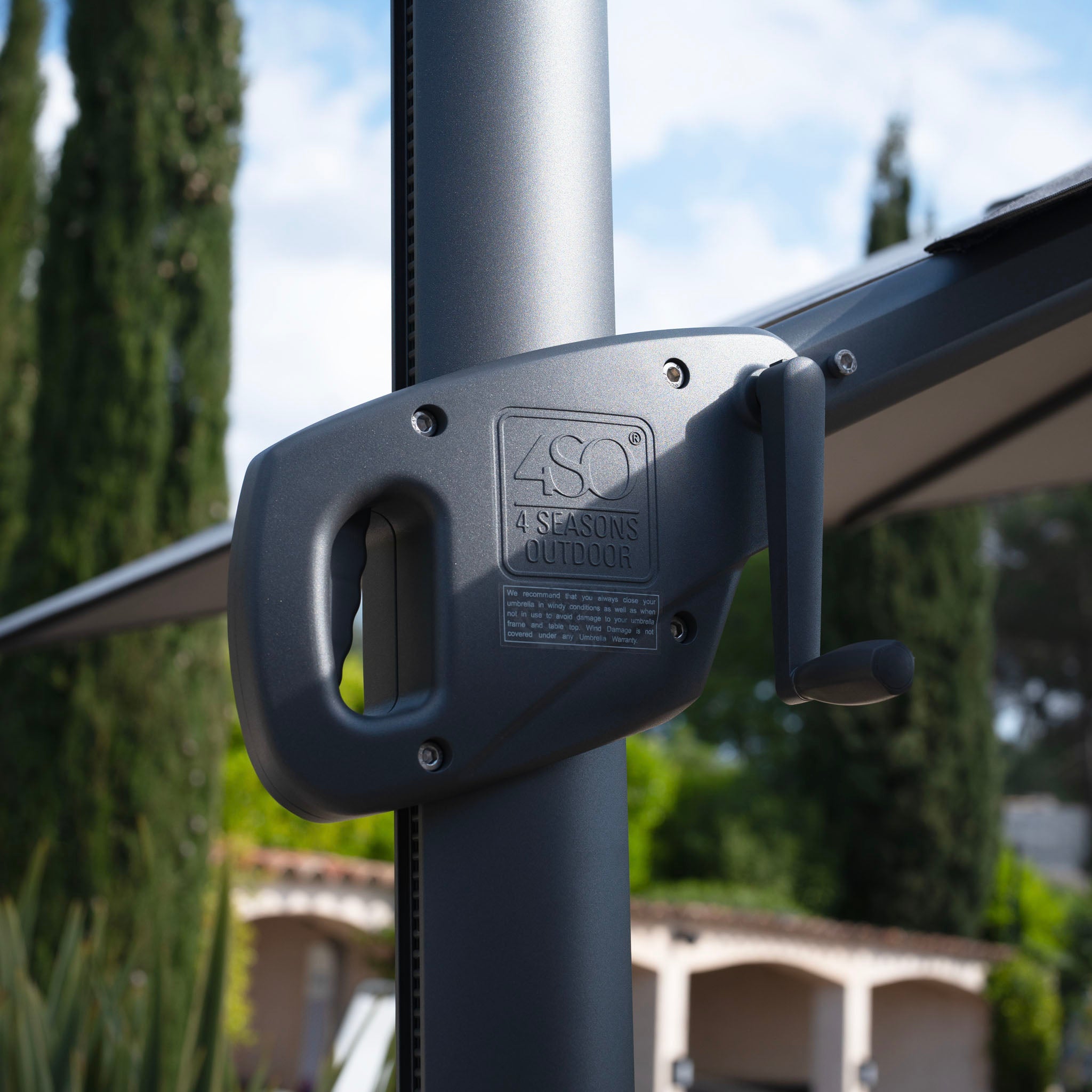 Close-up of the crank on a charcoal Hacienda 3m x 4m Cantilever Parasol with Granite Base & Cover, featuring "4 Seasons Outdoor" branding and highlighting its 360-degree rotation capability, set against a blurry background of trees and buildings.