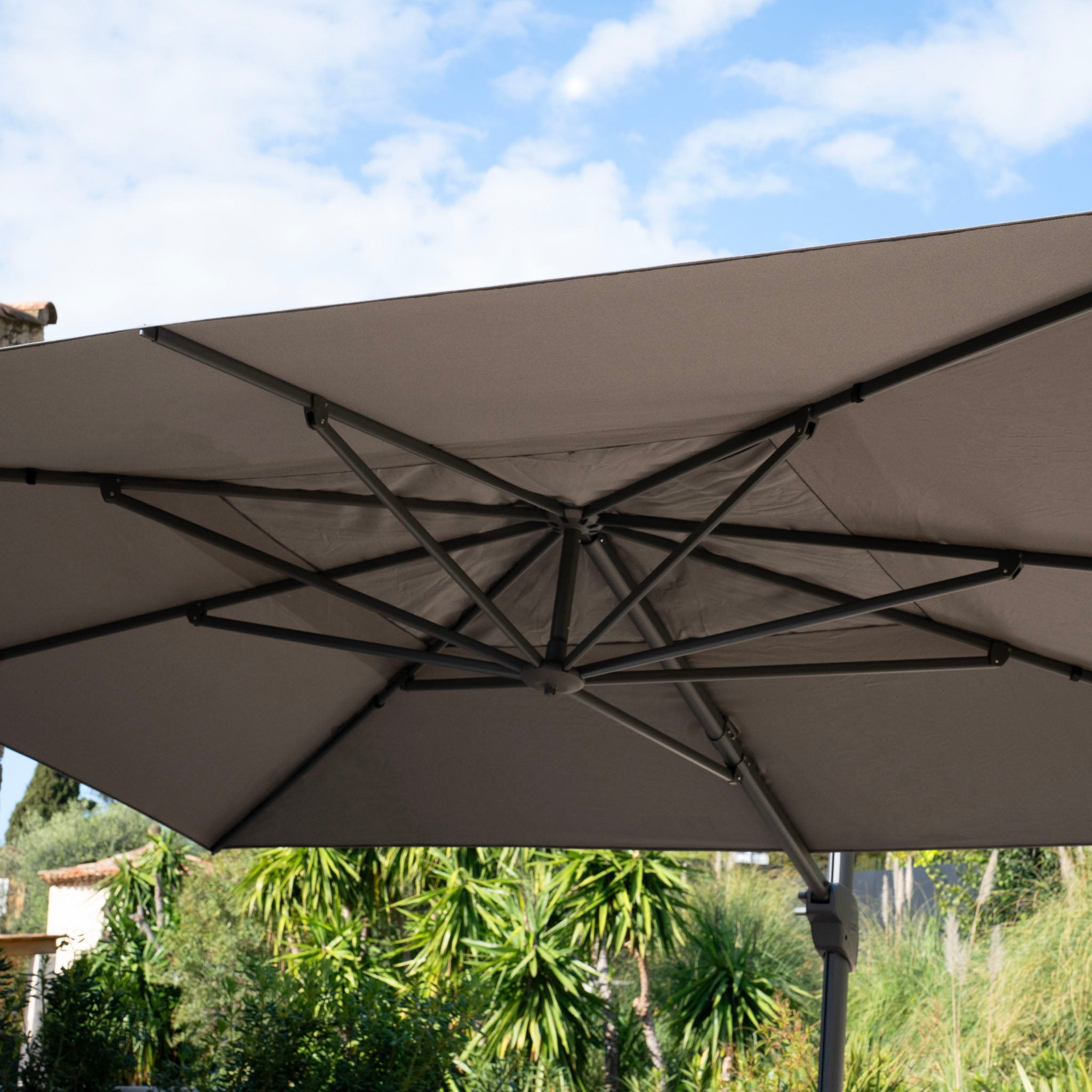 Close-up of the Hacienda 3m x 4m Cantilever Parasol with a granite base and cover in charcoal, showcasing its impressive UV-resistant canopy and sturdy metal frame against a clear sky and lush green foliage.