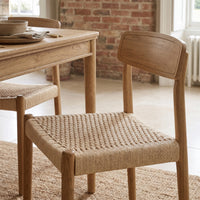 Hadley Set of 2 Dining Chairs in Natural with woven seats, paired with a wooden dining table in a sunlit room featuring brick and tile walls.