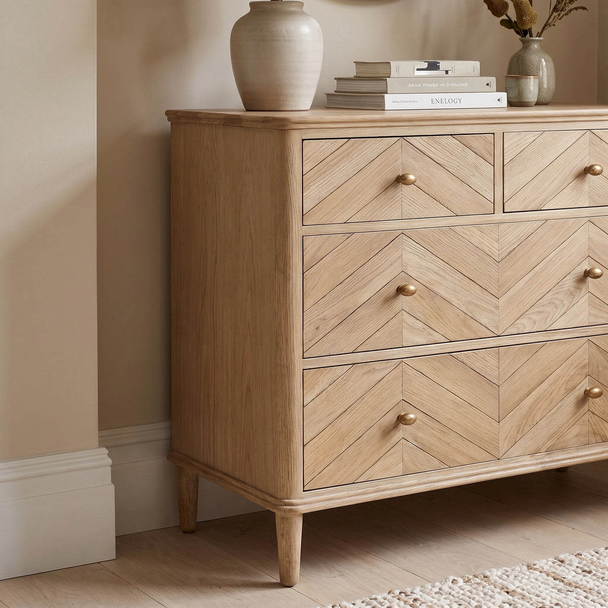 The Hadley Herringbone 2 Over 2 Chest of Drawers in Natural, featuring a chevron pattern and brass-effect knobs, is styled with books and a vase on top in a neutral room.