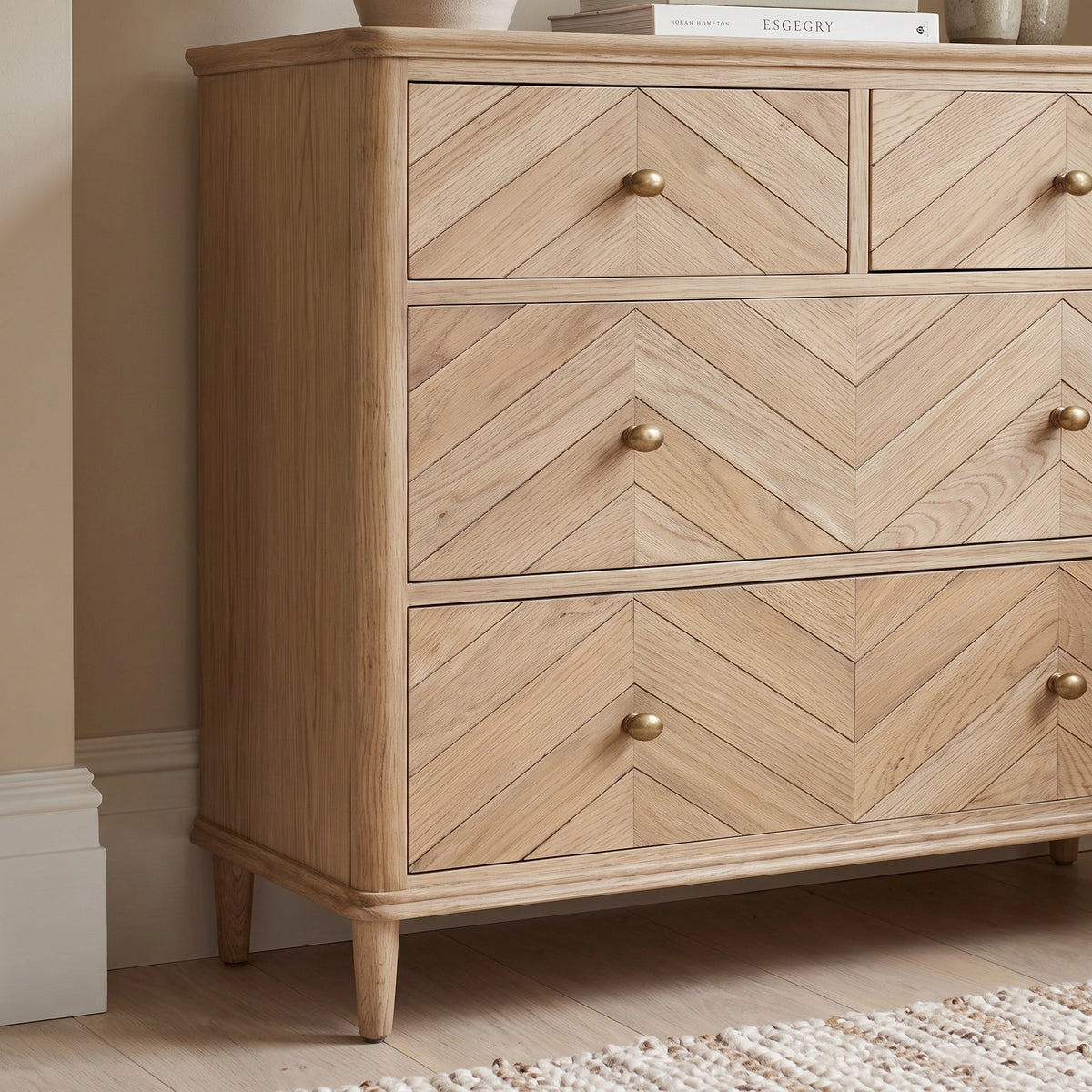 The Hadley Herringbone 2 Over 2 Chest of Drawers in Natural, featuring a chevron pattern, brass-effect knobs, and four barrel legs, sits on a rug in a neutral room.
