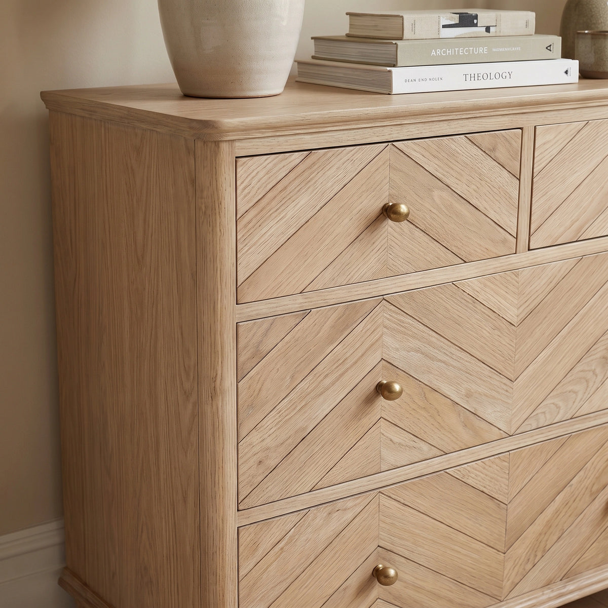 The Hadley Herringbone 2 Over 2 Chest of Drawers in Natural features light wood and chevron pattern drawers with brass-effect knobs, topped with books and a vase.