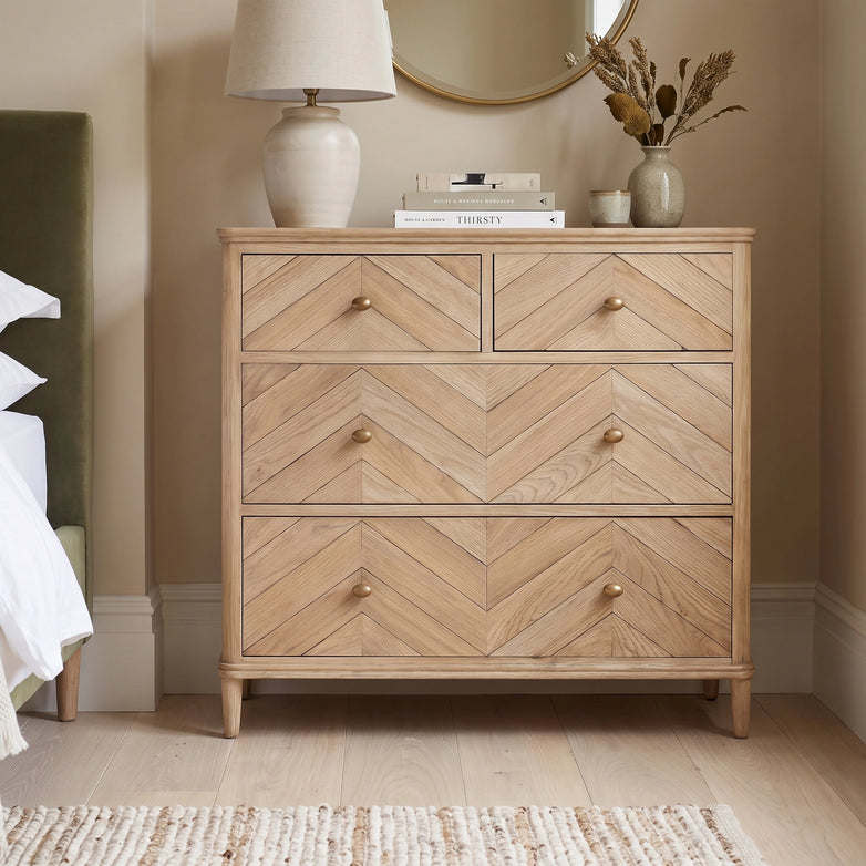 The Hadley Herringbone 2 Over 2 Chest of Drawers in Natural, styled with a lamp, books, and a vase on top, sits next to a bed in a cozy room.