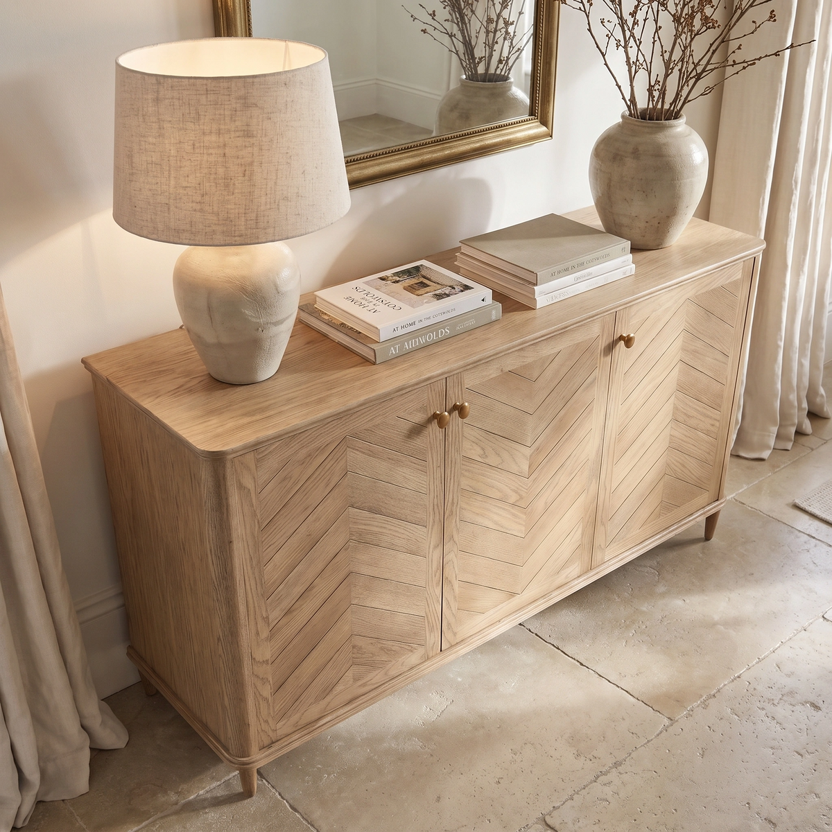 The Hadley Herringbone 3 Door Sideboard in Natural, featuring a chevron pattern, sits beneath a mirror and is adorned with a lamp, books, and a vase.