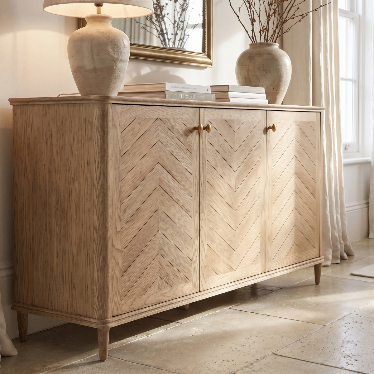 Hadley Herringbone 3 Door Sideboard in Natural with chevron pattern, styled with two vases, books, and a lamp on top and a mirror above.