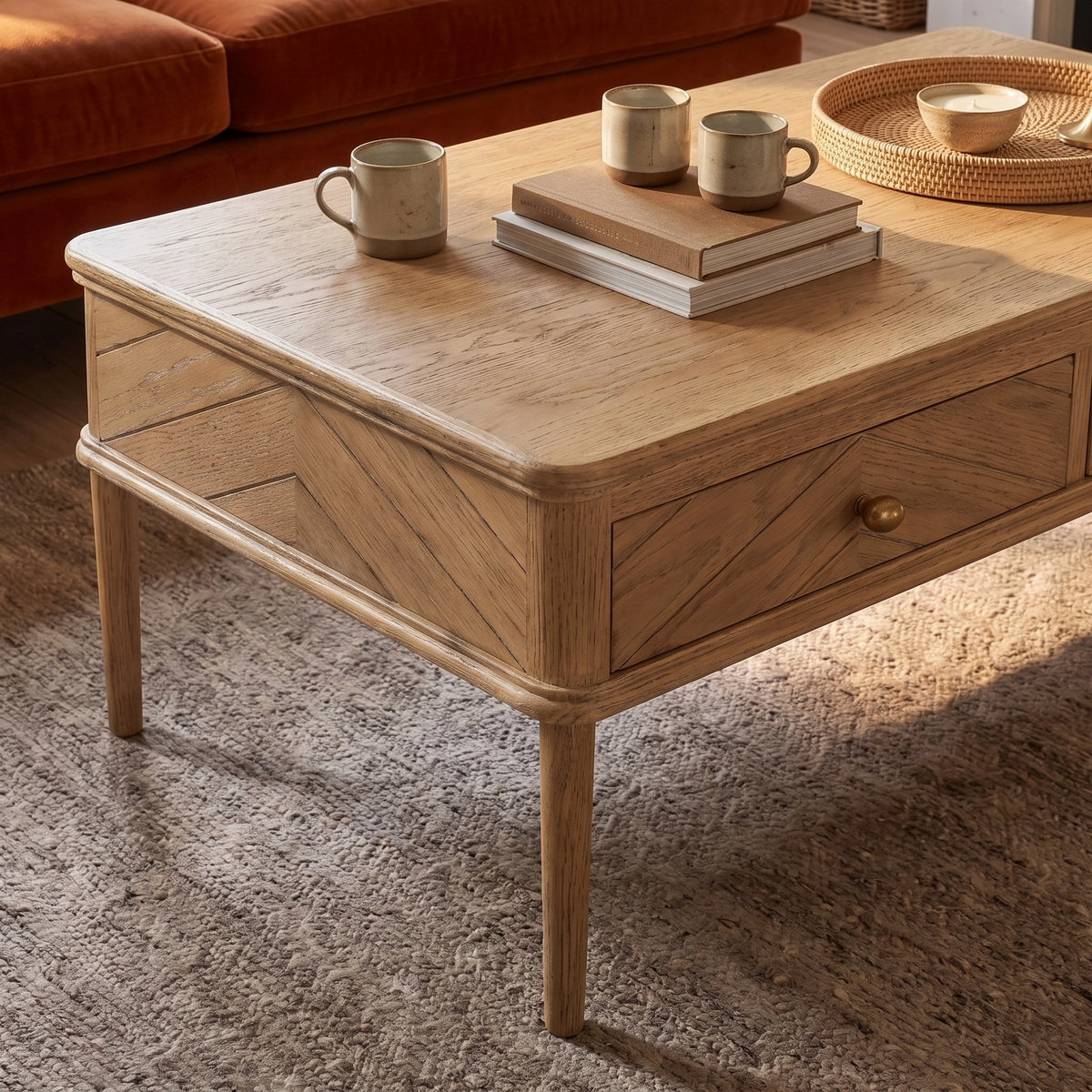 Placed atop a textured rug beside an orange sofa, the Hadley Herringbone Coffee Table in Natural brings understated sophistication to the room. This chevron parquet table showcases its light wood finish while displaying three mugs, two books, and a tray—making it an inviting and stylish focal point for any living area.
