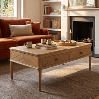 A Hadley Herringbone Coffee Table in Natural, adorned with mugs and books, sits in a cozy living room furnished with a sofa and fireplace.