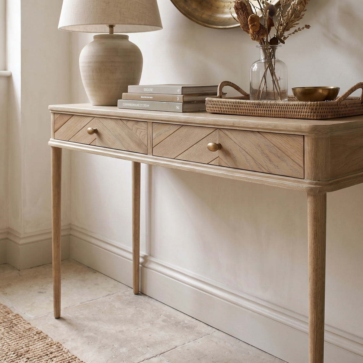 Hadley Herringbone Console Table in Natural with two drawers, styled with stacked books, a lamp, vase, and decor in a neutral-toned room.