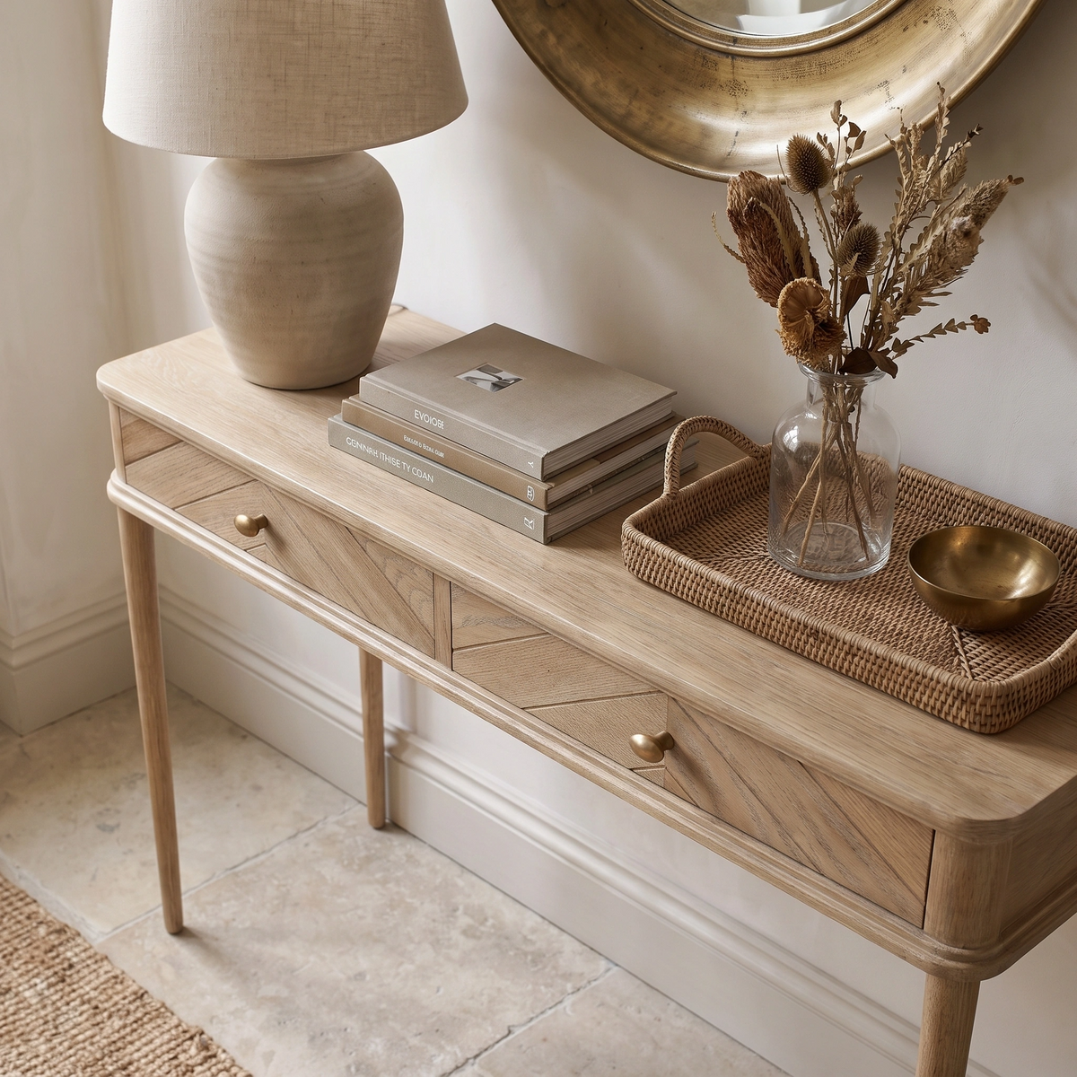 Hadley Herringbone Console Table in Natural styled with a lamp, books, dried flowers in a jar, and a round mirror above.