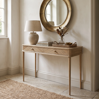 A Hadley Herringbone Console Table in Natural is styled with a lamp, books, a bowl, and dried flowers beneath a round mirror.