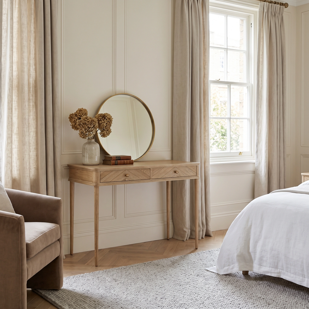 Light, airy bedroom with beige curtains, a Hadley Herringbone Console Table in Natural, a round mirror, and dried flowers in a vase.
