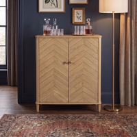 Hadley Herringbone Drinks Cabinet in Natural, featuring a chevron pattern and light oak finish, styled with decanters on top and placed beside a floor lamp and framed wall art.