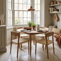 The Hadley Herringbone 4-6 Round Extending Dining Table in Natural is paired with four chairs, creating a cozy, sunlit kitchen nook by large bay windows.