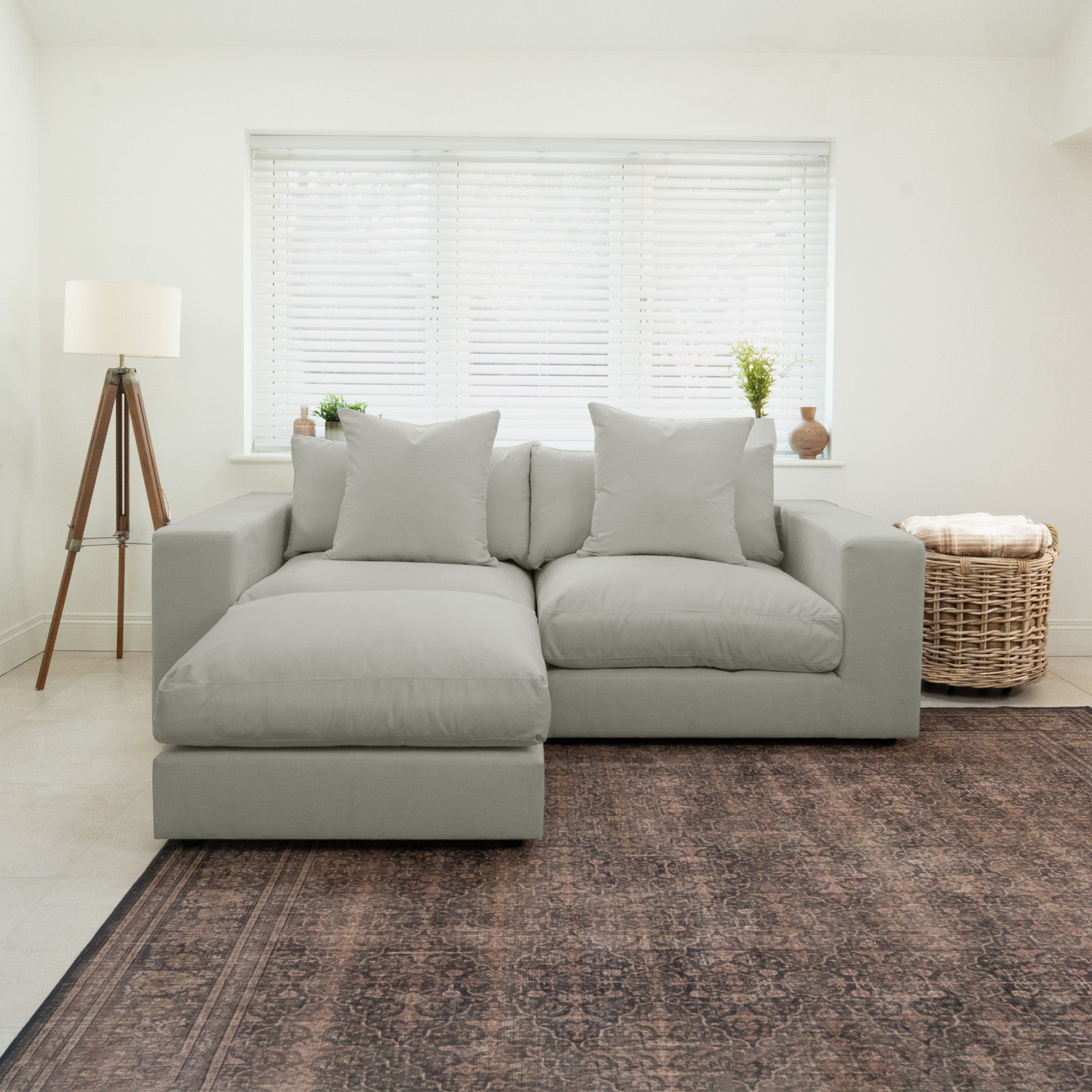 The Hampshire Modular 2 Seat Sofa in Classic Grey, featuring duck feather and down cushions, sits in a bright living room with a lamp and wicker basket nearby.