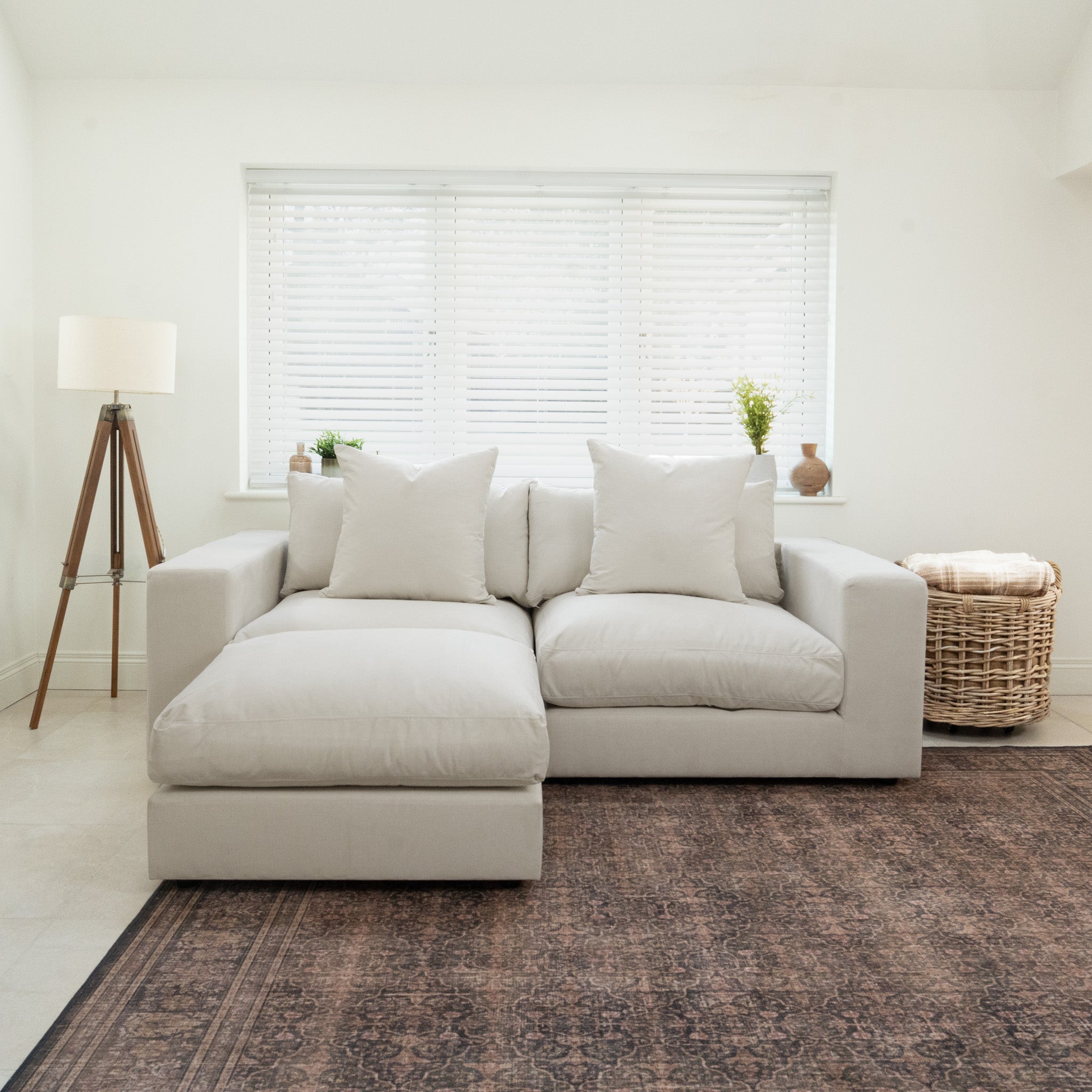 The Hampshire Modular 2 Seat Sofa in Warm Grey sits in a bright living room with a floor lamp, potted plants, and a woven basket. Crafted from sustainably sourced hardwood, it features luxurious duck feather and down cushions.