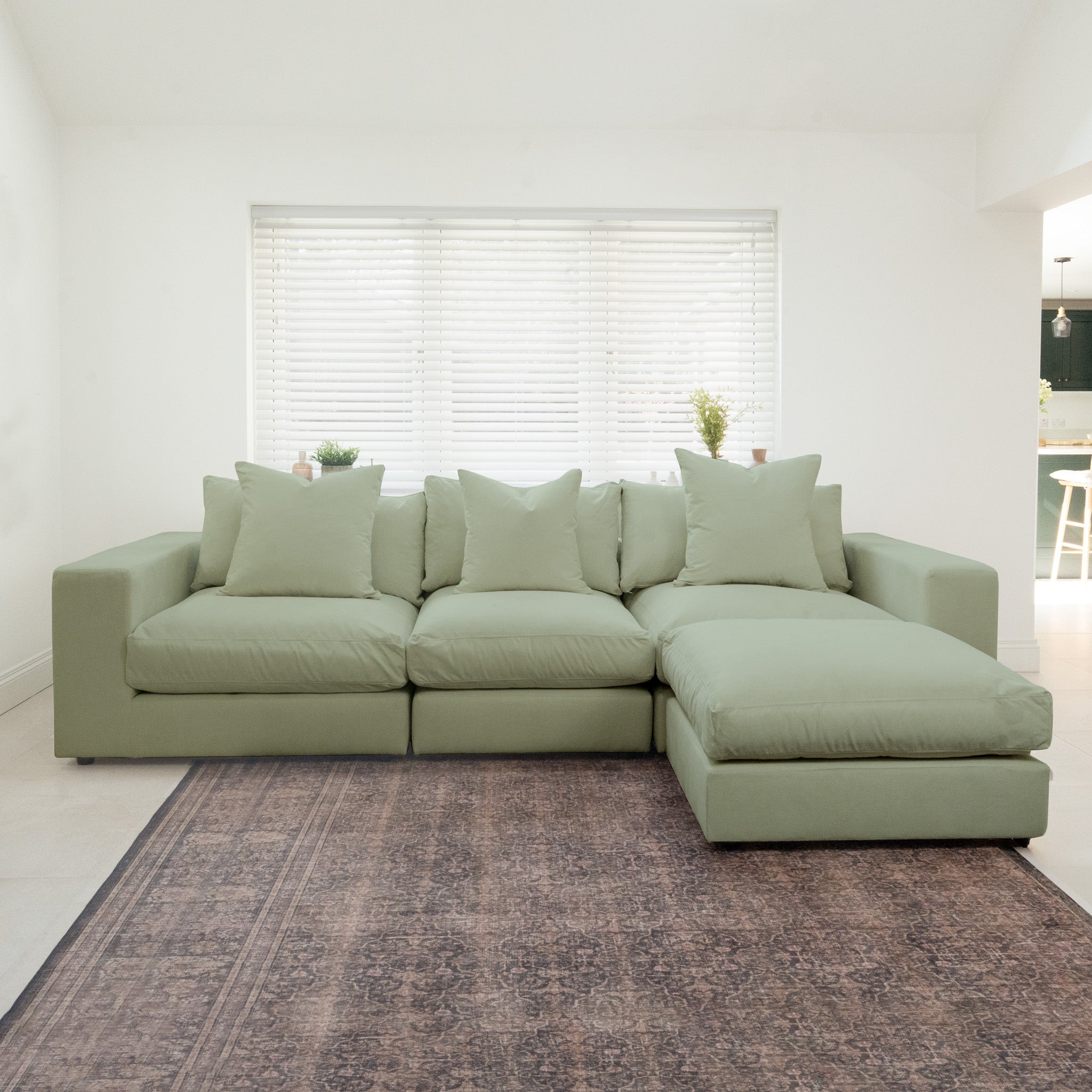 The Hampshire Modular 3 Seat Sofa in Moss Green with duck feather cushions rests on a patterned rug in a bright room with large window blinds.