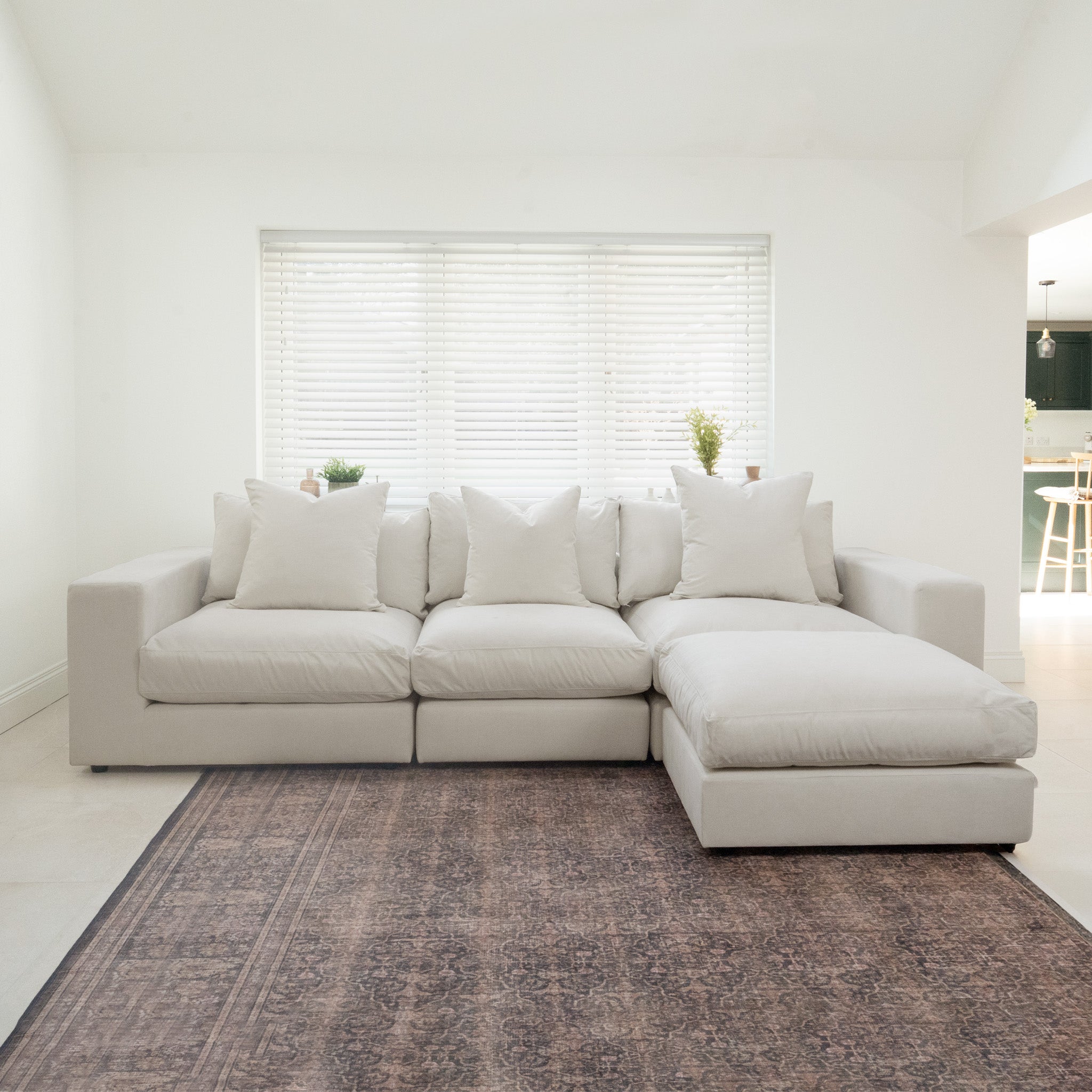The Hampshire Modular 3 Seat Sofa in Warm Grey, crafted from sustainably sourced hardwood with plush duck feather cushions, sits atop a patterned rug in a bright room with a window and blinds.