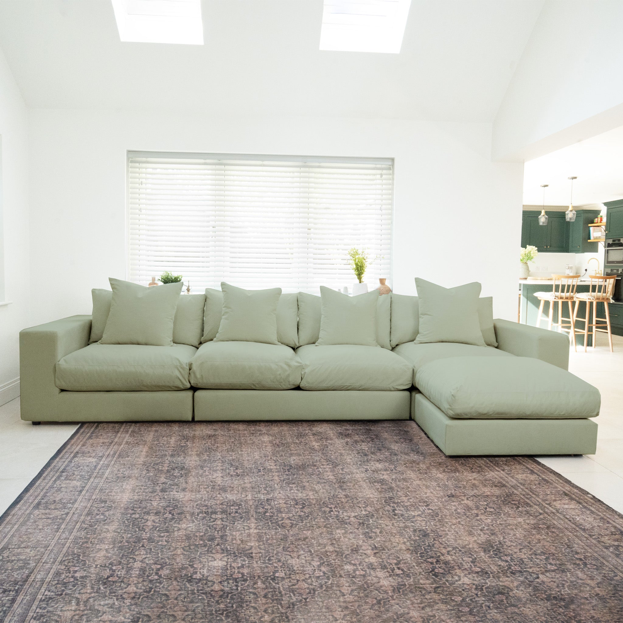 The Hampshire Modular 4 Seat Sofa in Moss Green, made from sustainably sourced hardwood and topped with five plush duck feather and down cushions, sits on a patterned rug in a bright, modern living room.