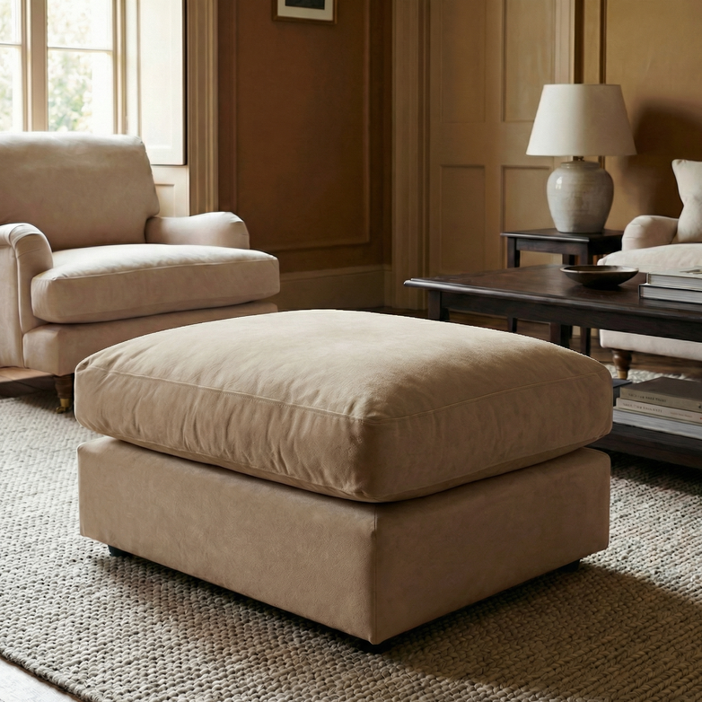 The Hampshire Modular Footstool in Beige, handmade in England with linen-effect fabric, brings charm to a cozy living room when paired with a matching armchair, rug, lamp, and wooden coffee table.