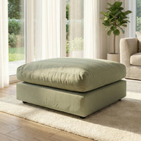 A Hampshire Modular Footstool in moss green sits on a cream rug in a bright living room with large windows and a potted plant.