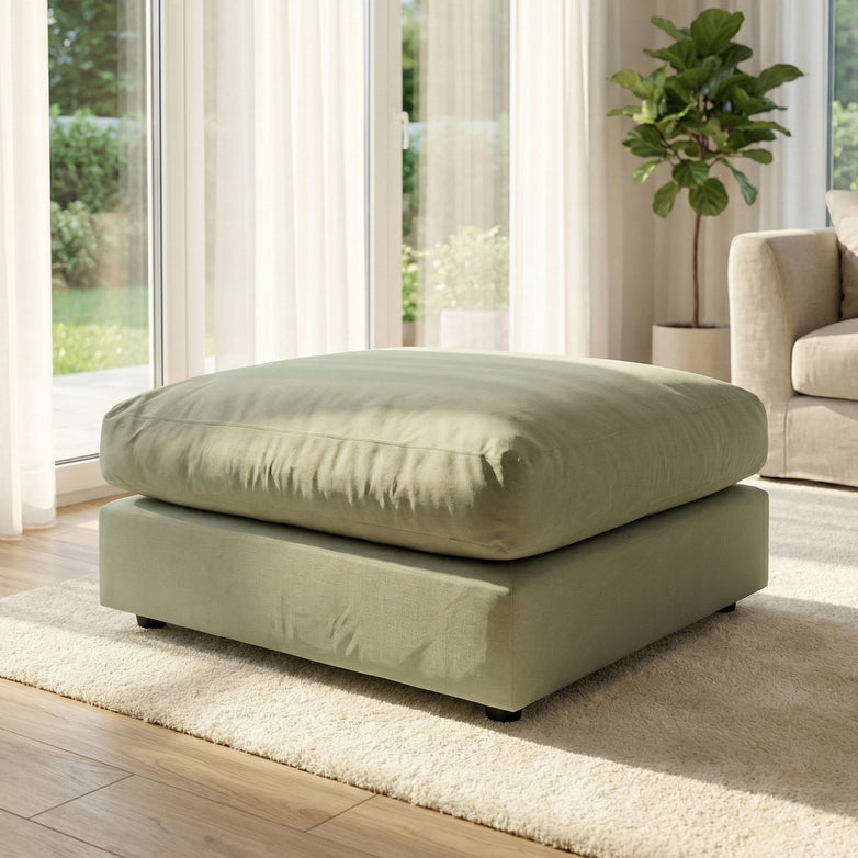 A Hampshire Modular Footstool in moss green sits on a cream rug in a bright living room with large windows and a potted plant.