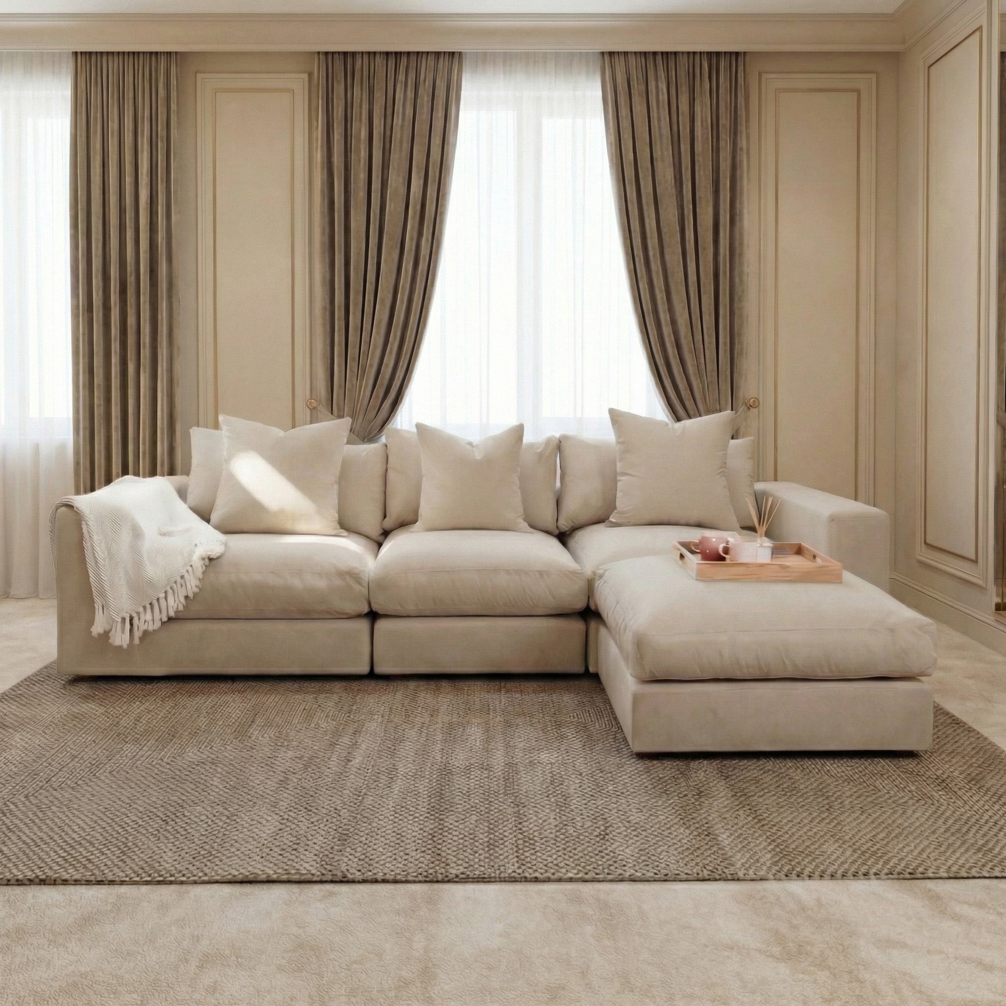 The Hampshire Modular 3 Seat Sofa in Beige features duck feather cushions, a cozy throw blanket, and a tray, creating an elegant living room setting with large windows and floor-length curtains.
