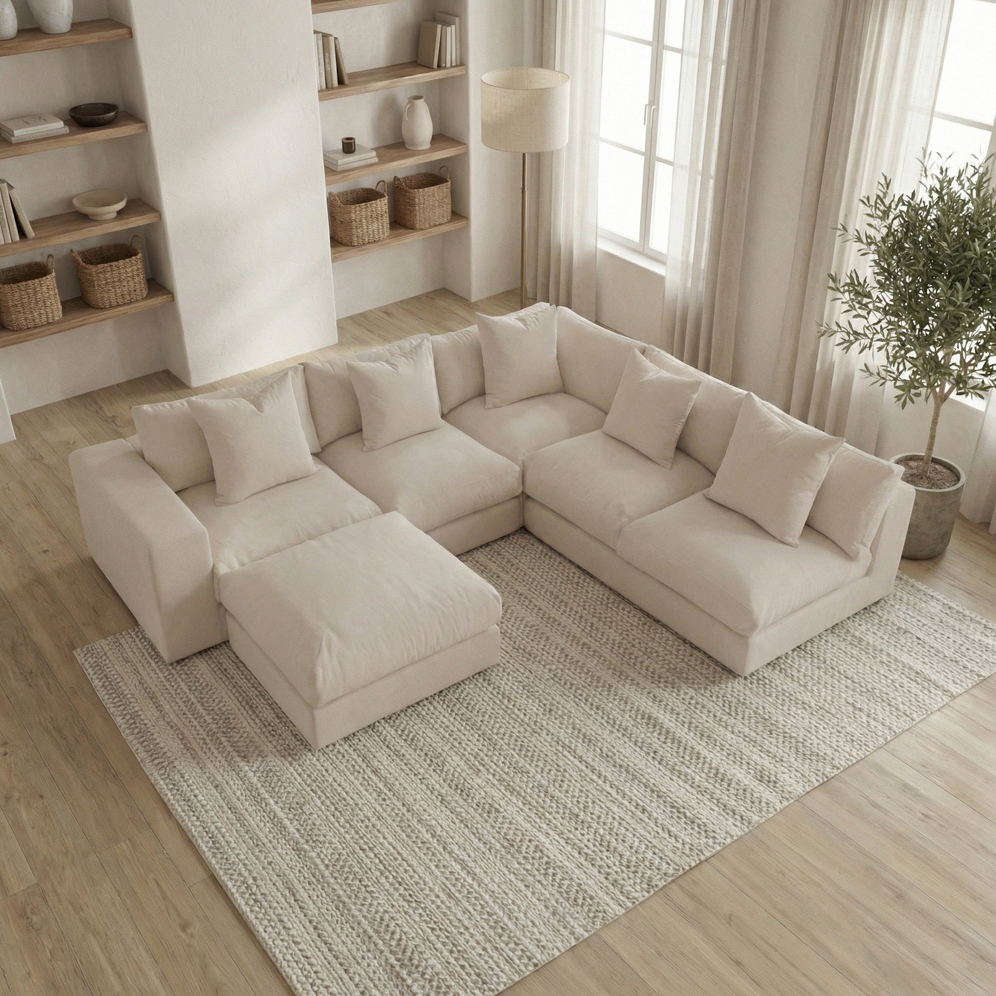 The Hampshire Modular Corner Sofa in Beige with matching ottoman sits on a textured rug in a bright, modern living room with shelves and plants; handmade in England for timeless comfort and style.