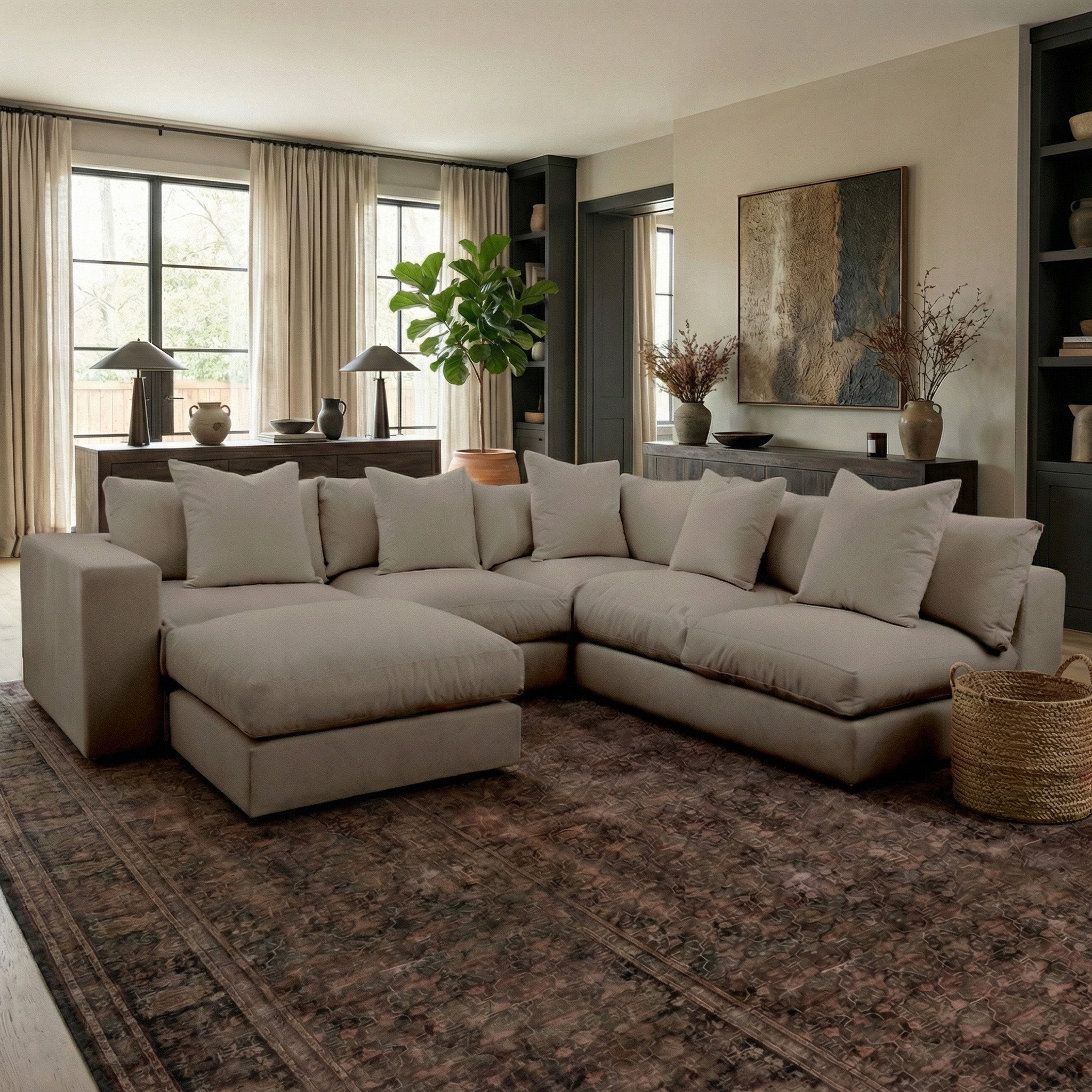 A Hampshire Modular Corner Sofa in Cocoa, handmade in England and piled with pillows, sits on a patterned rug in a softly lit modern living room.