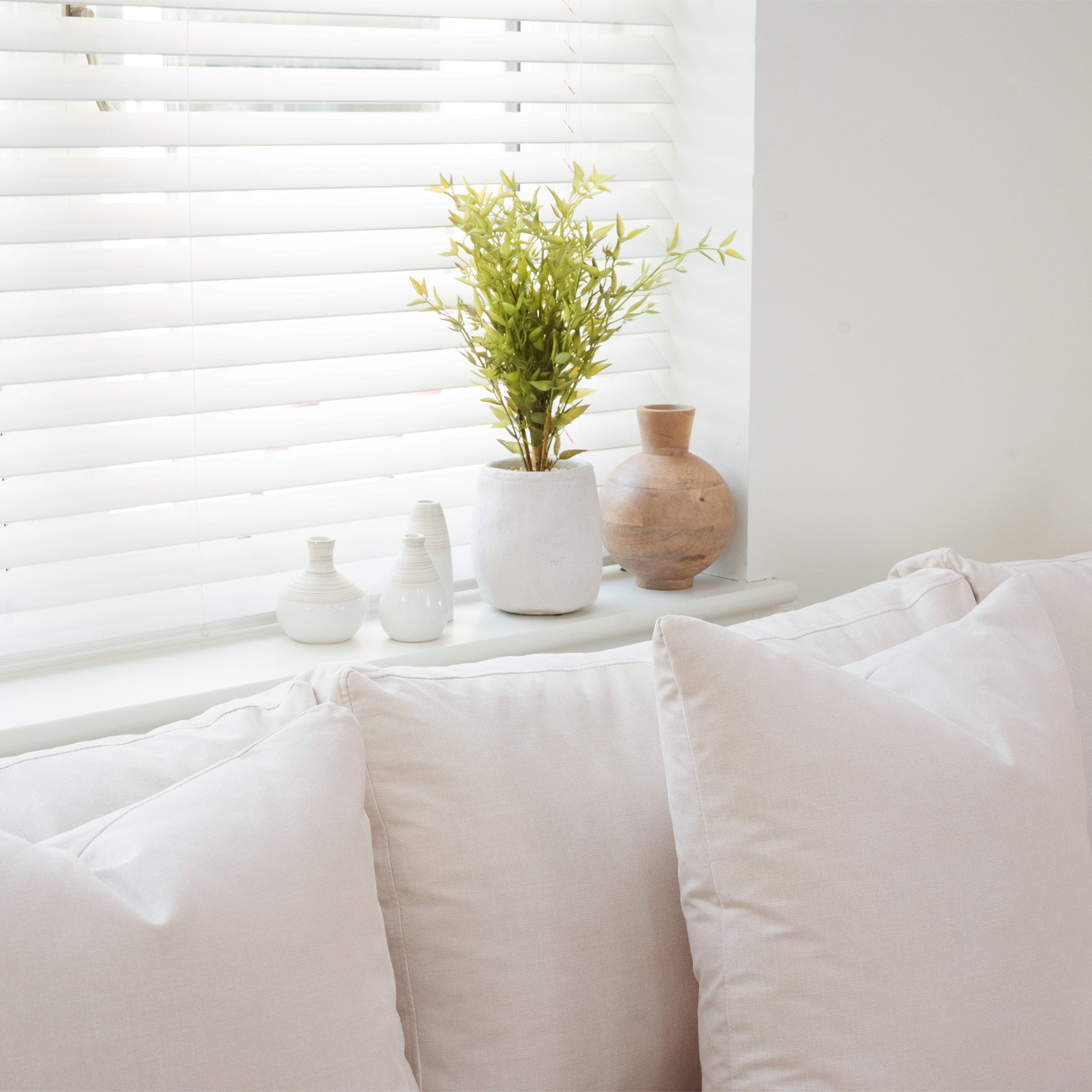 The Hampshire Modular Corner Sofa in Ivory features luxurious duck feather and down cushions, sustainably sourced hardwood, and sits elegantly by a window with blinds, a potted plant, and ceramic vases on the sill for eco-friendly comfort.