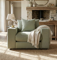 A cozy Hampshire Modular Armchair in Moss Green, handmade in England with plush duck feather and down cushions, sits with pillows, a throw blanket, and a tea cup in a sunlit living room.