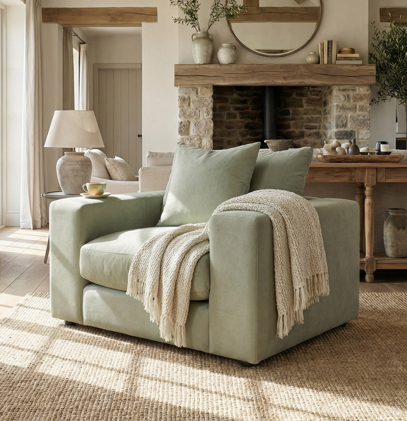 A Hampshire Modular Armchair in Moss Green with duck feather cushions and a throw blanket sits in a sunlit living room, surrounded by rustic decor and a stone fireplace.