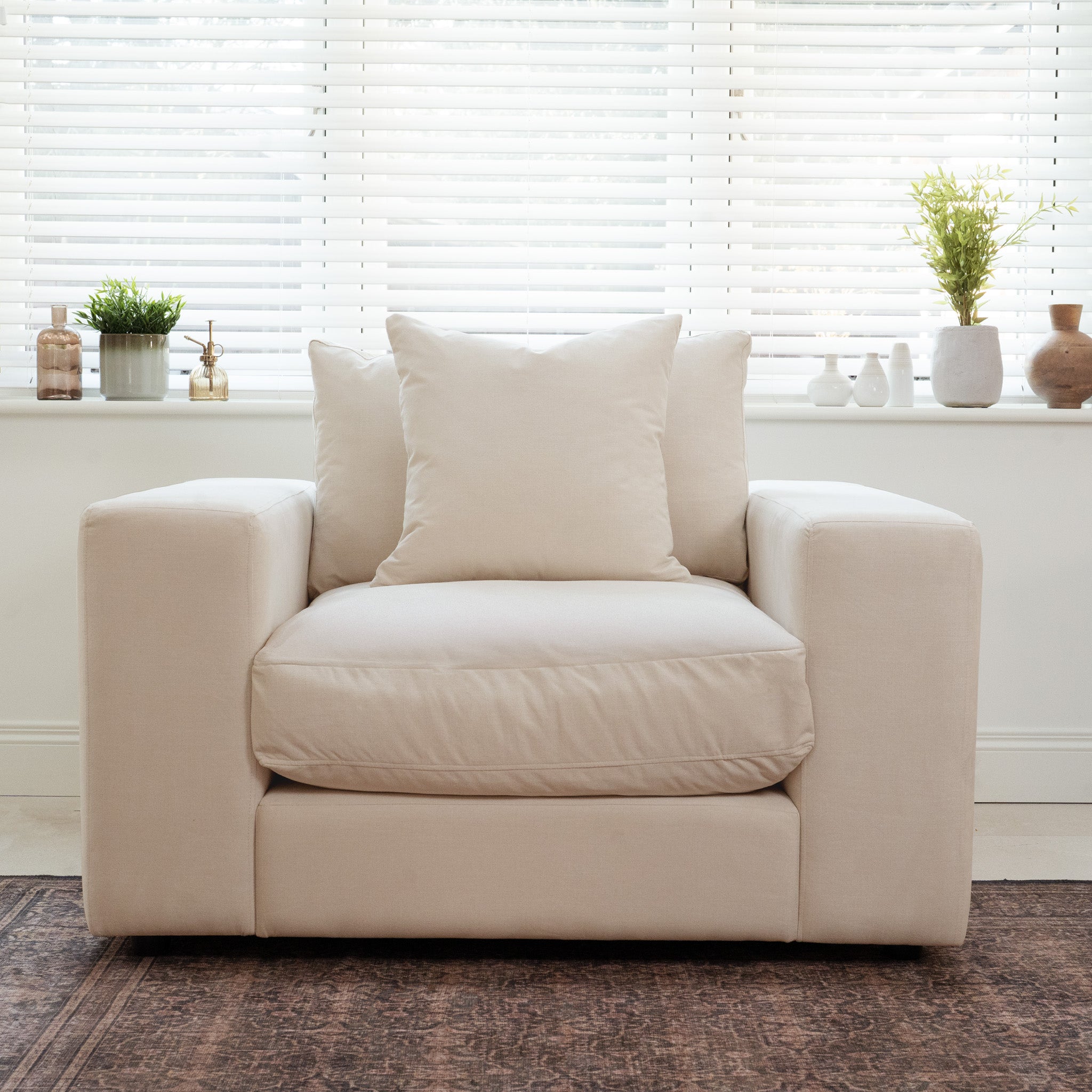 The Hampshire Modular Armchair in Beige with duck feather cushions is placed in front of a window with blinds and potted plants on the sill.