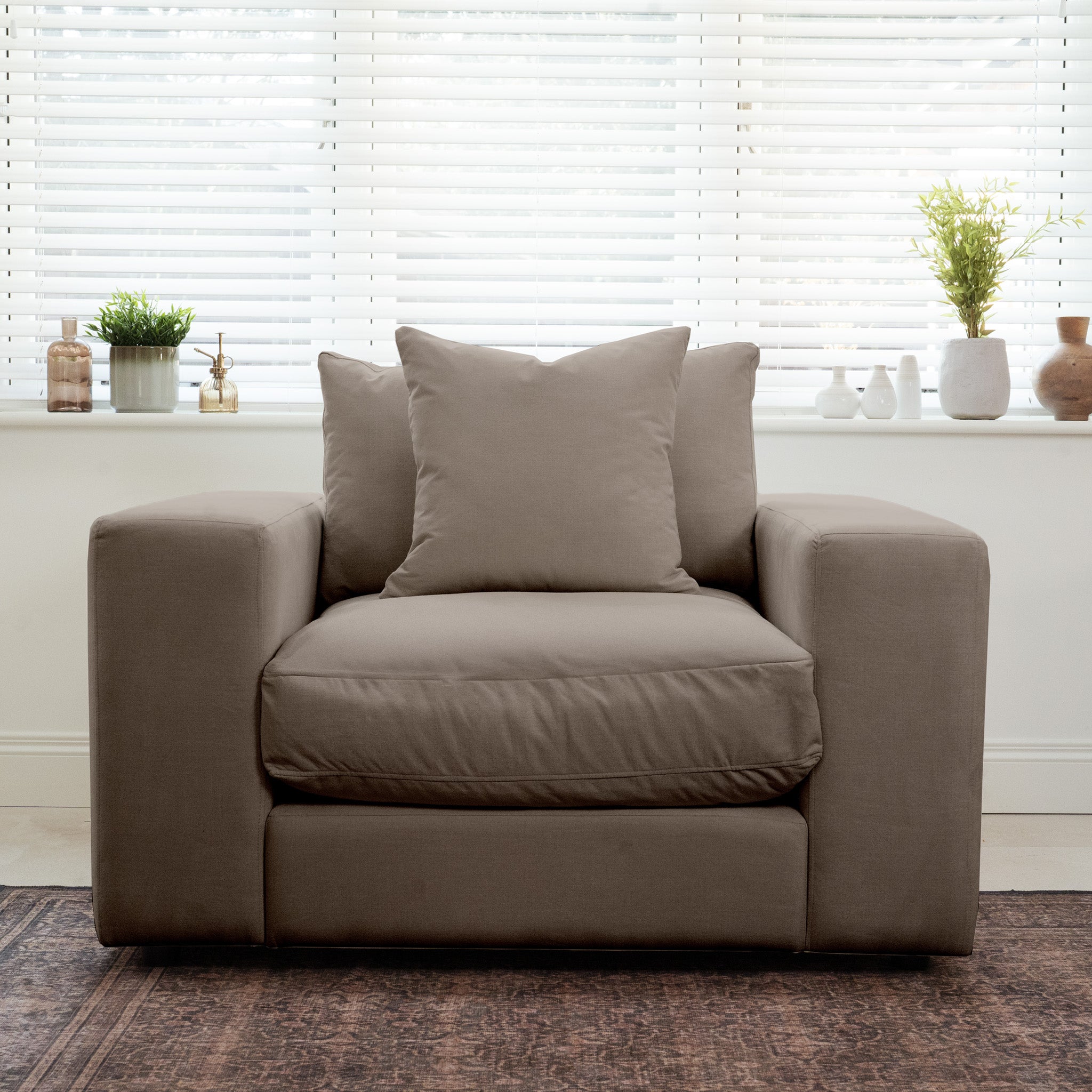 A spacious cocoa Hampshire Modular Armchair with duck feather and down cushions sits in front of a window, accented by pillows and potted plants on the windowsill.