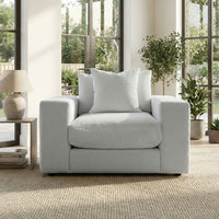 The Hampshire Modular Armchair in Dove Grey, handmade in England with plush duck feather cushions and white pillows, sits in a bright room with large windows and green plants.