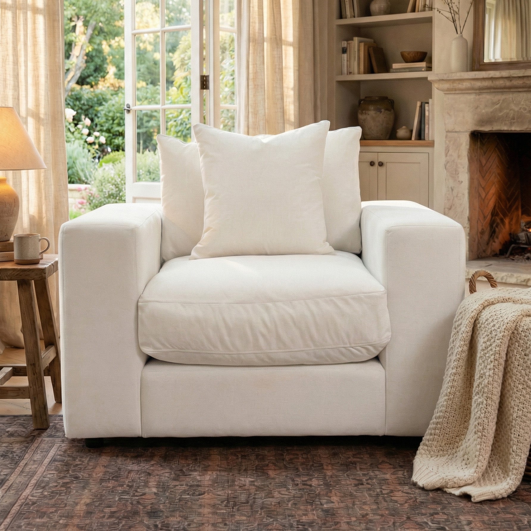 The Hampshire Modular Armchair in Ivory, handmade in England with duck feather cushions and a knitted blanket, sits in a cozy living room filled with natural light and views of the garden.