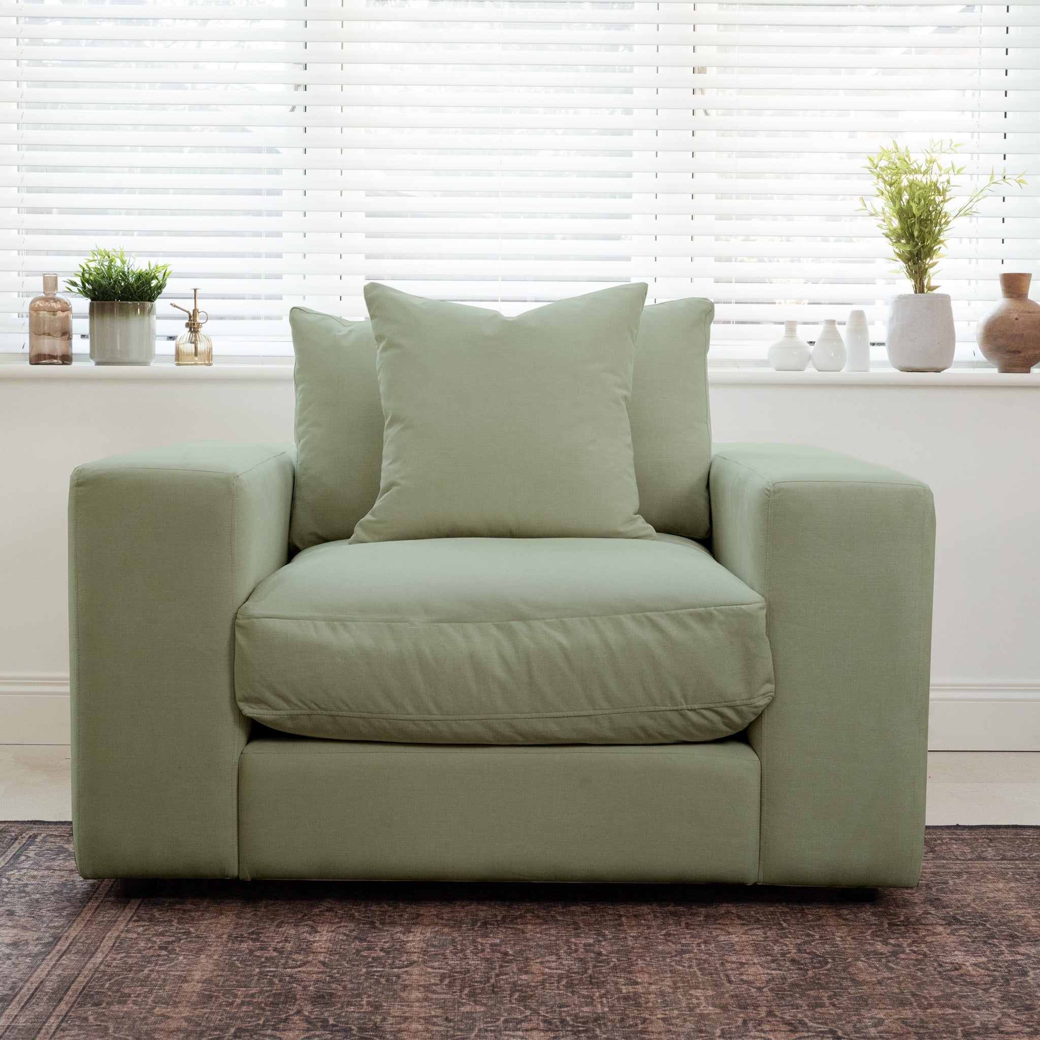 The Hampshire Modular Armchair in Moss Green, crafted from sustainably sourced hardwood and featuring luxurious cushions, sits before a window with blinds and potted plants on the sill.
