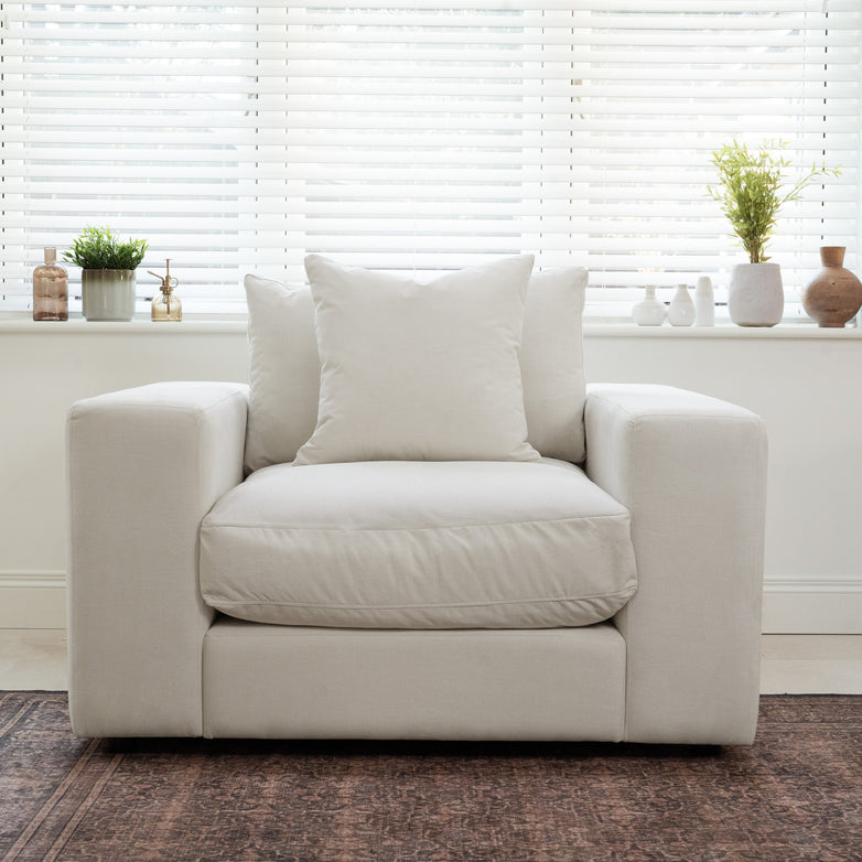 The Hampshire Modular Armchair in Warm Grey, crafted from sustainably sourced hardwood with plush cushions, is positioned by a window with blinds and potted plants.