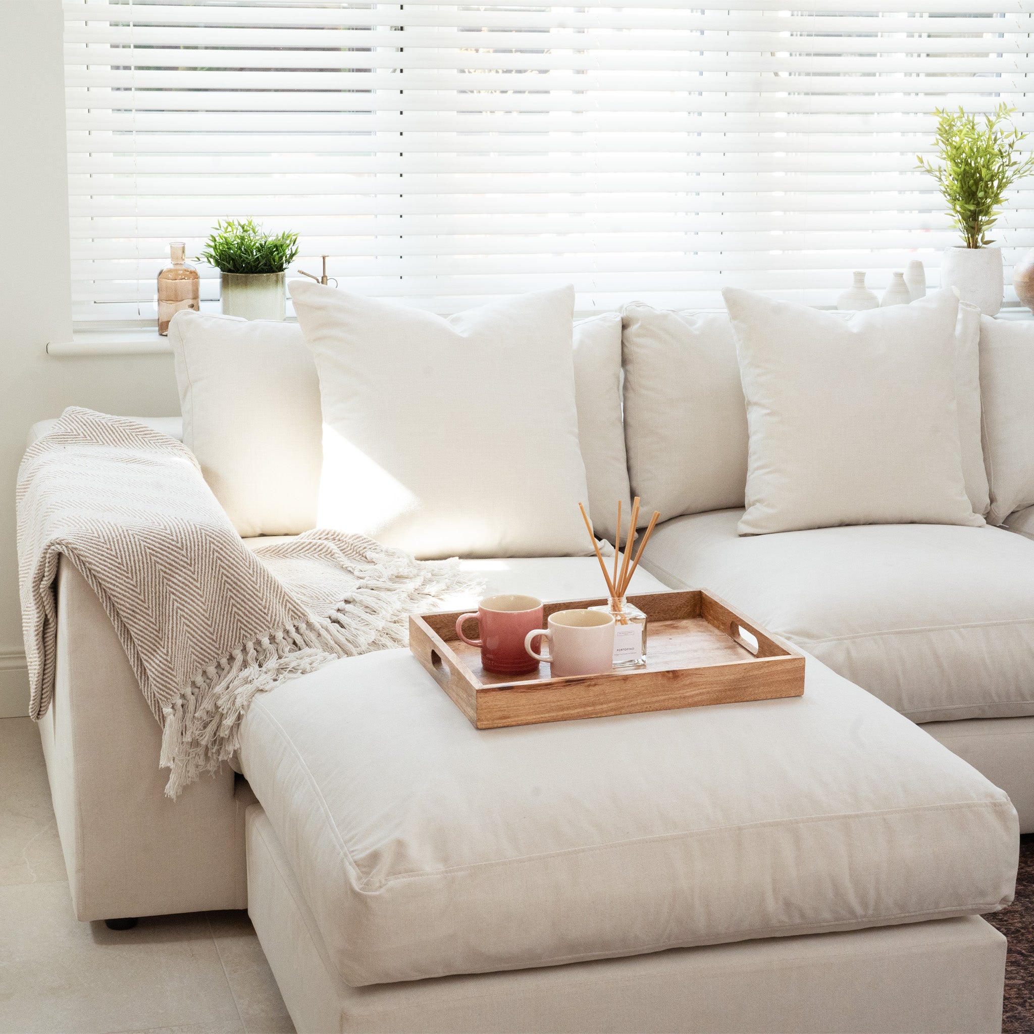 Hampshire Modular Corner Sofa in Ivory with white pillows, a throw blanket, and a wooden tray holding mugs in a bright living room—crafted from sustainably sourced hardwood for lasting elegance.