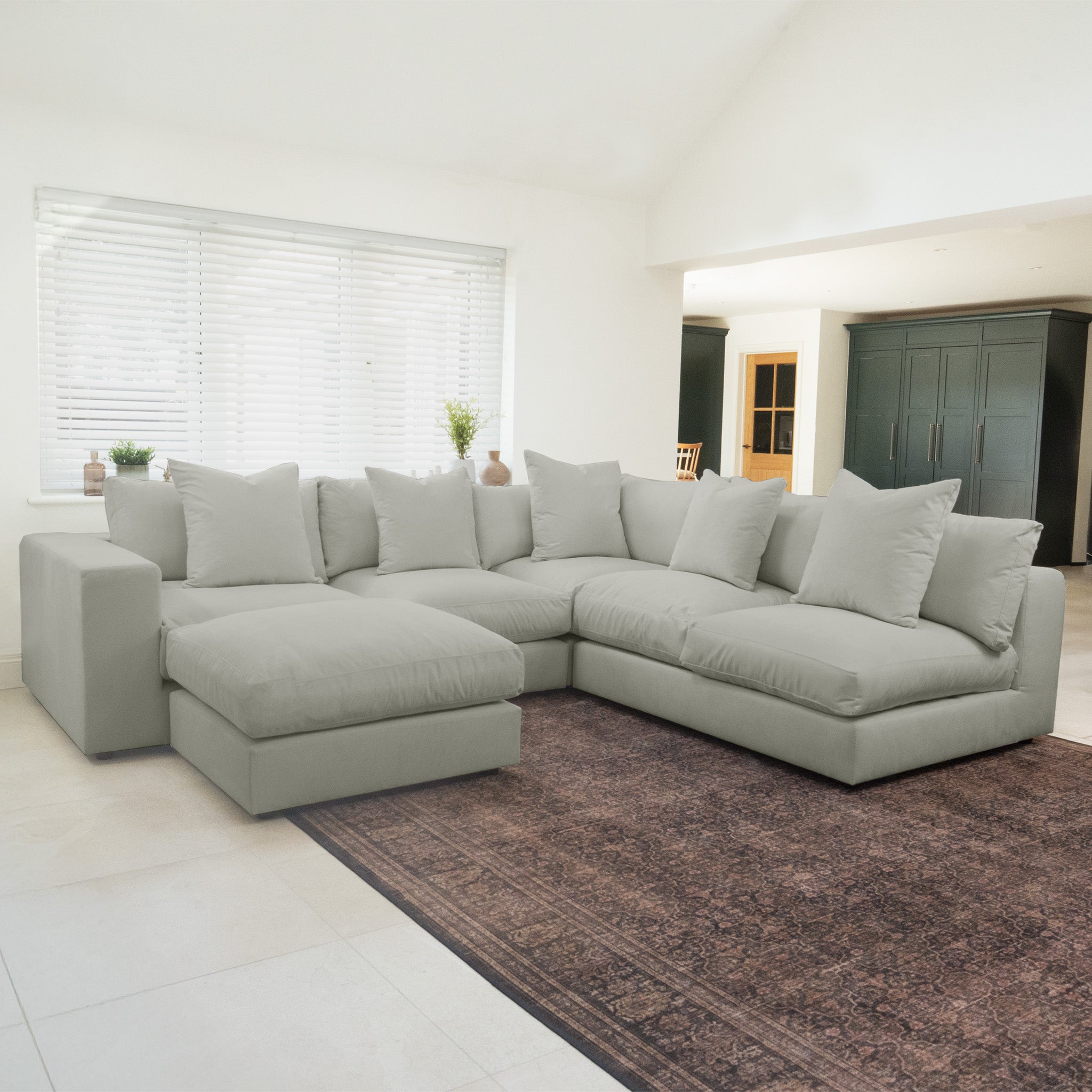 The Hampshire Modular Corner Sofa in Classic Grey, featuring duck feather and down cushions, sits on a patterned brown rug in a bright modern living room.