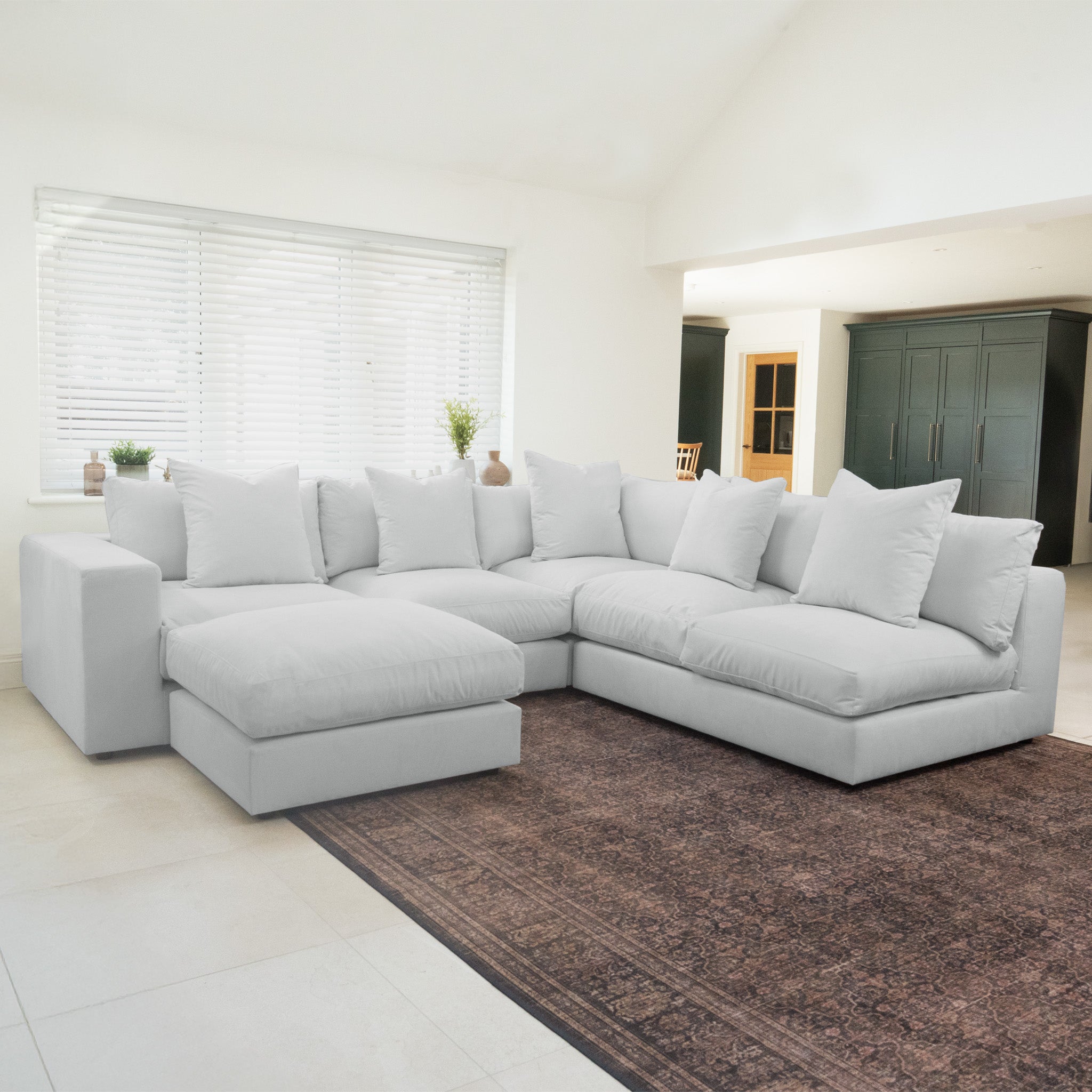 A spacious Hampshire Modular Corner Sofa in Dove Grey with plush duck feather cushions sits on a brown rug in a bright, modern living room.