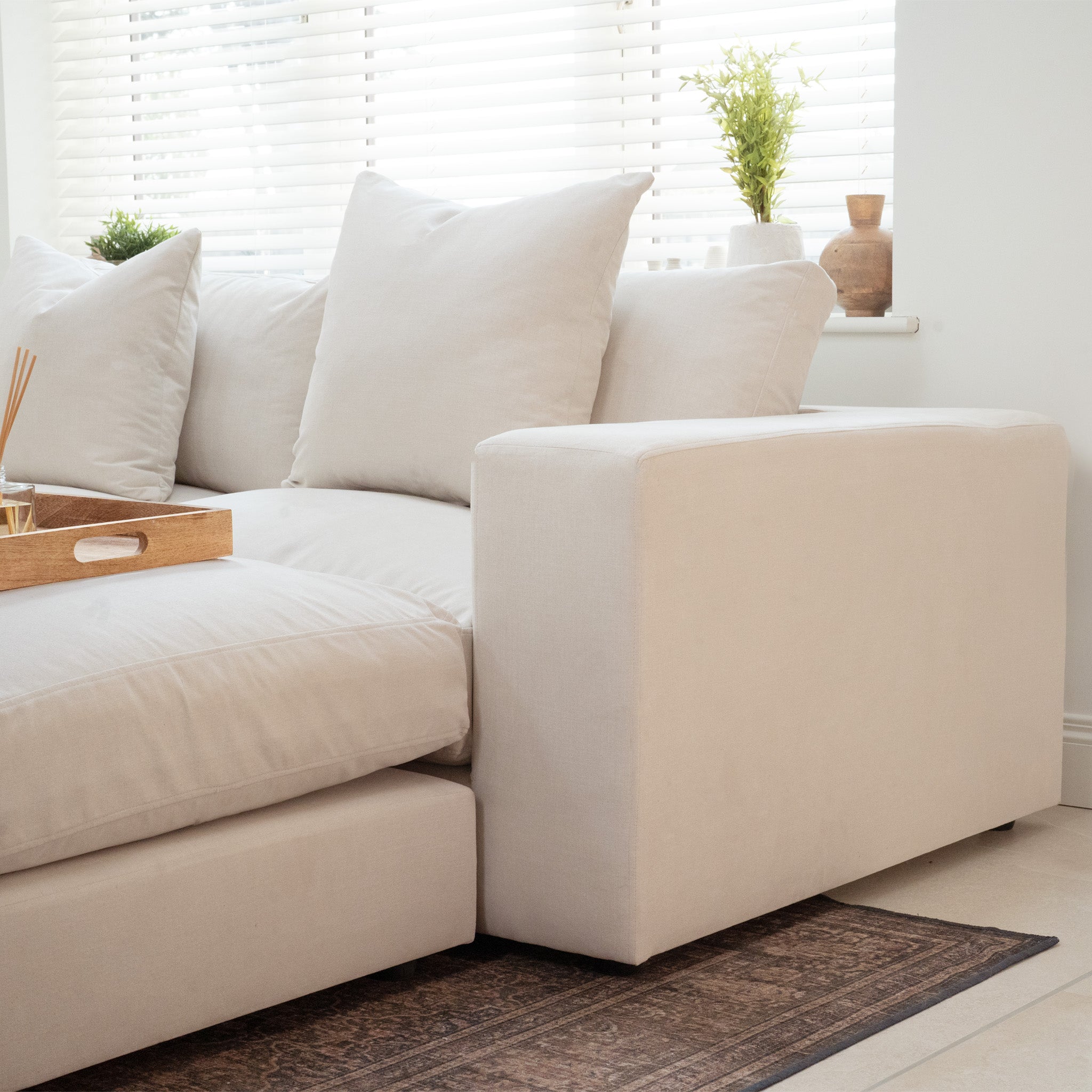 The Hampshire Modular Corner Sofa in Ivory, crafted from sustainably sourced hardwood with duck feather and down cushions, is placed by a window with blinds and potted plants.