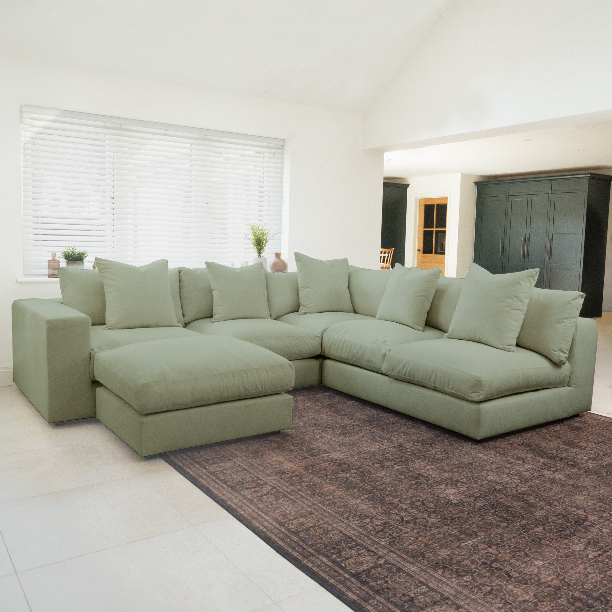 The Hampshire Modular Corner Sofa in Moss Green with duck feather cushions sits atop a large dark rug in a bright, modern living room and is crafted from sustainably sourced hardwood.
