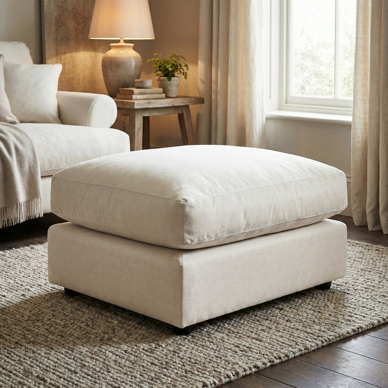 The Hampshire Modular Footstool in Ivory sits on a textured rug in a cozy living room with a sofa, side table, and window.