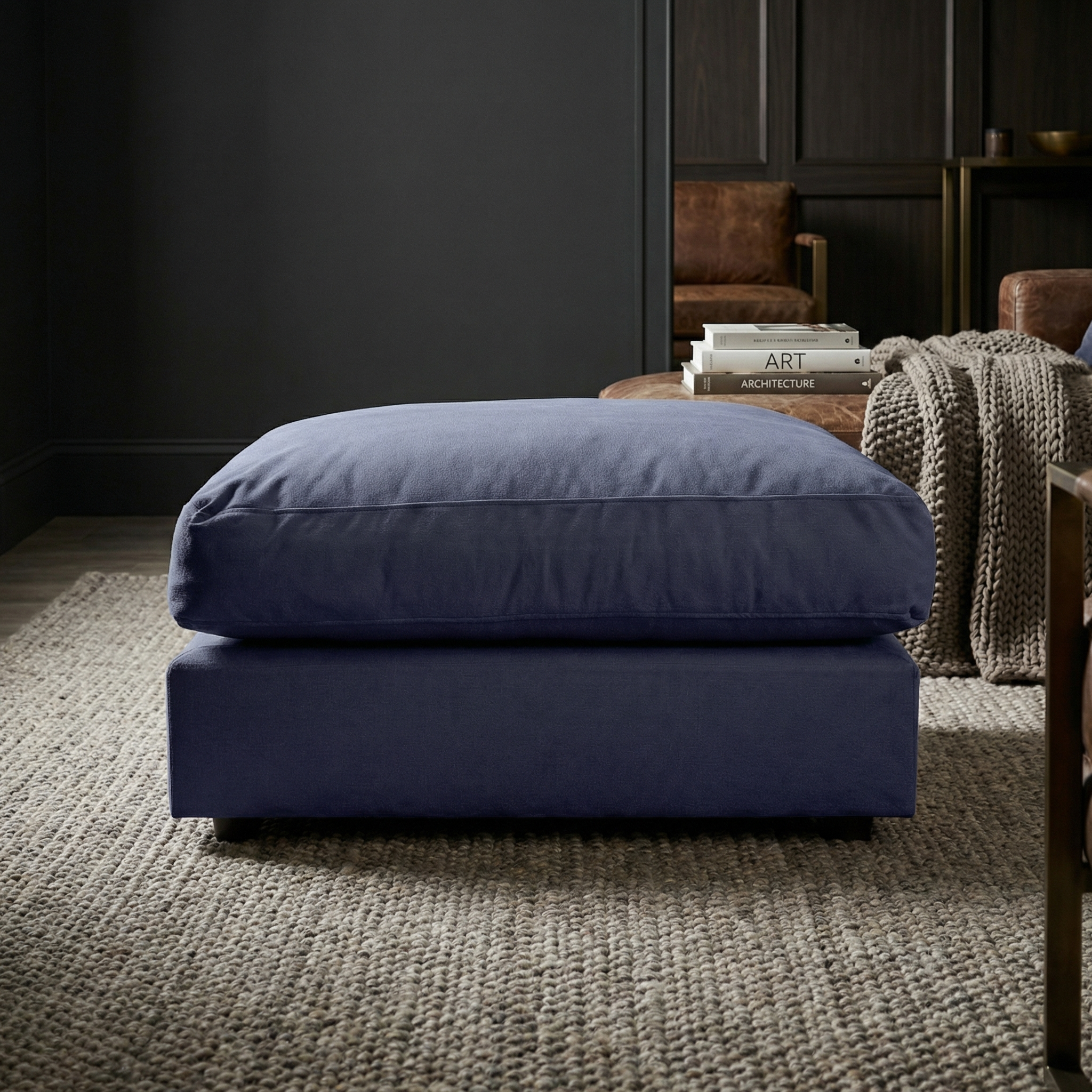 The Hampshire Modular Footstool in Midnight Blue sits on a textured rug in a modern living room with dark walls and cozy decor; handmade in England.