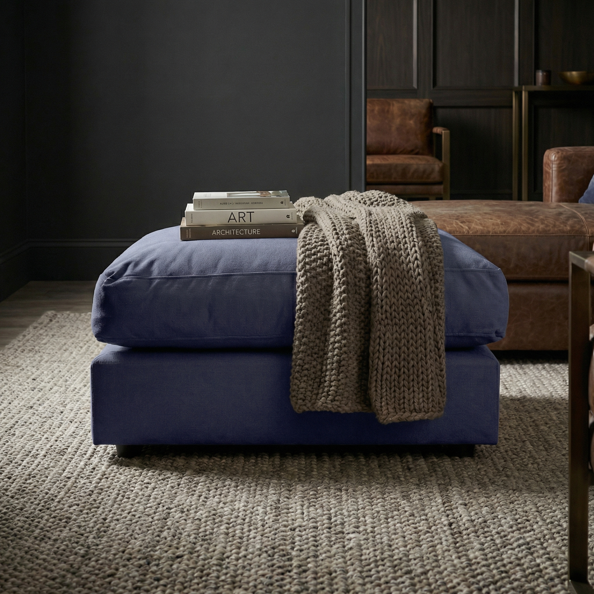 A Hampshire Modular Footstool in Midnight Blue, paired with books and a knit blanket, rests on a textured rug in a cozy, modern living room.